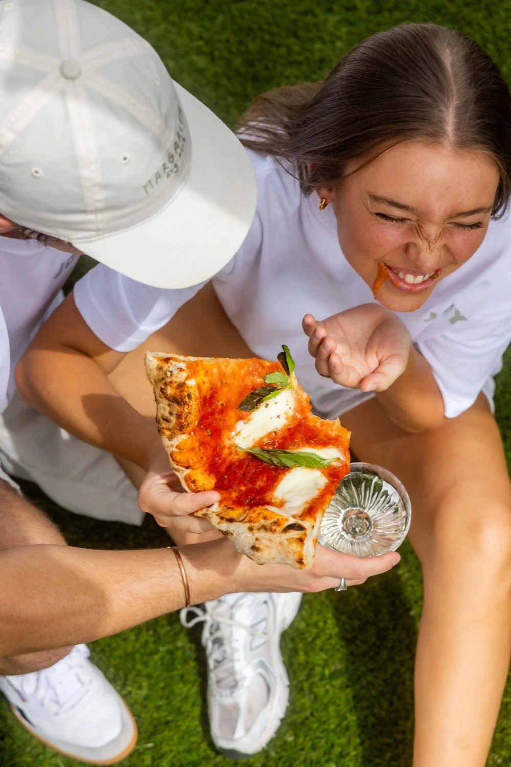 A girl with long hair laughing as she receives a slice of pepperoni pizza in a glass dish from a person wearing a white hat. They are outdoors on green grass.