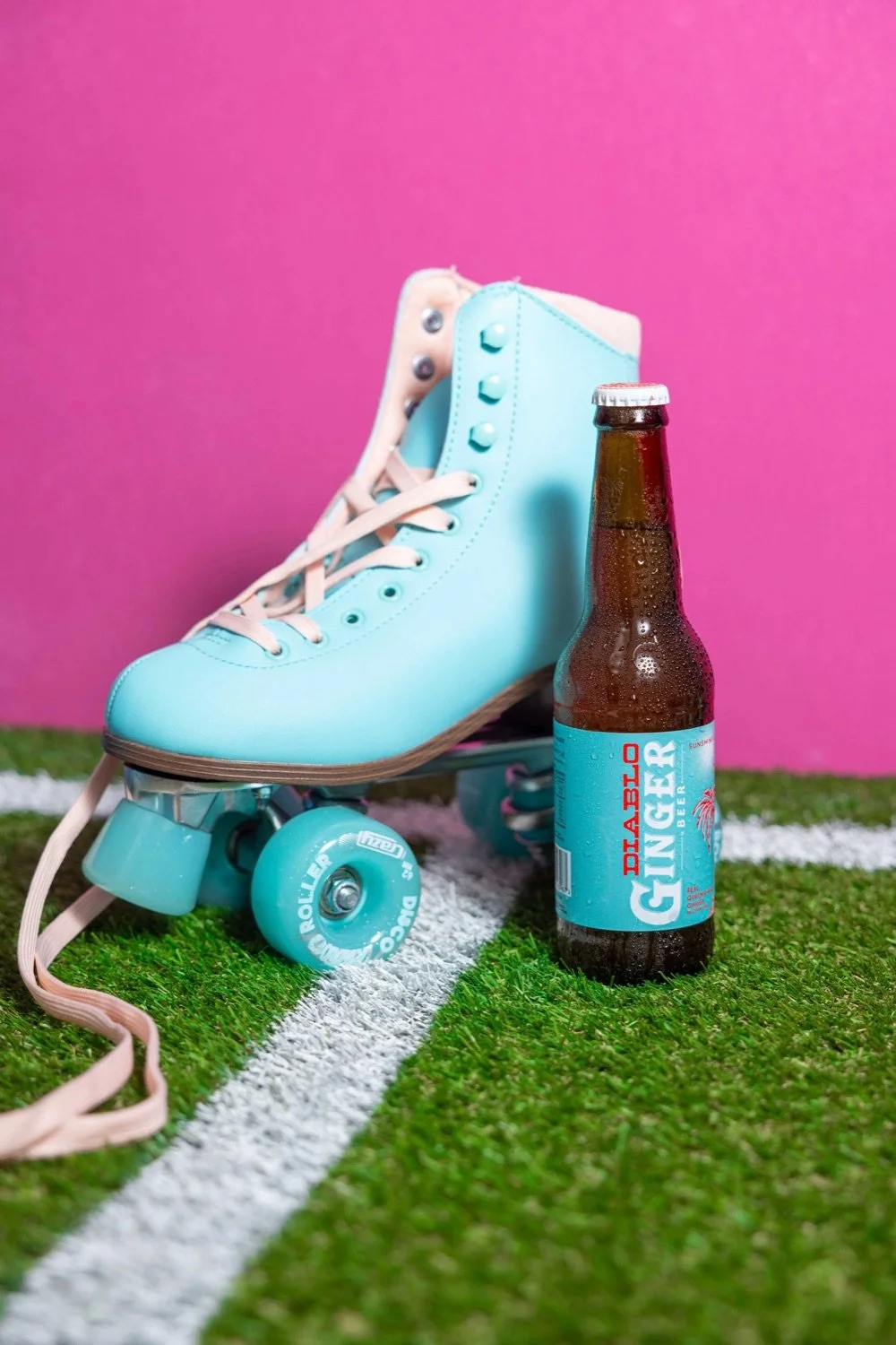 A pair of light blue roller skates, a bottle of ginger beer labeled 'Diablo Ginger Beer', and light blue roller skate wheels on artificial grass with white lane markings and a pink background.