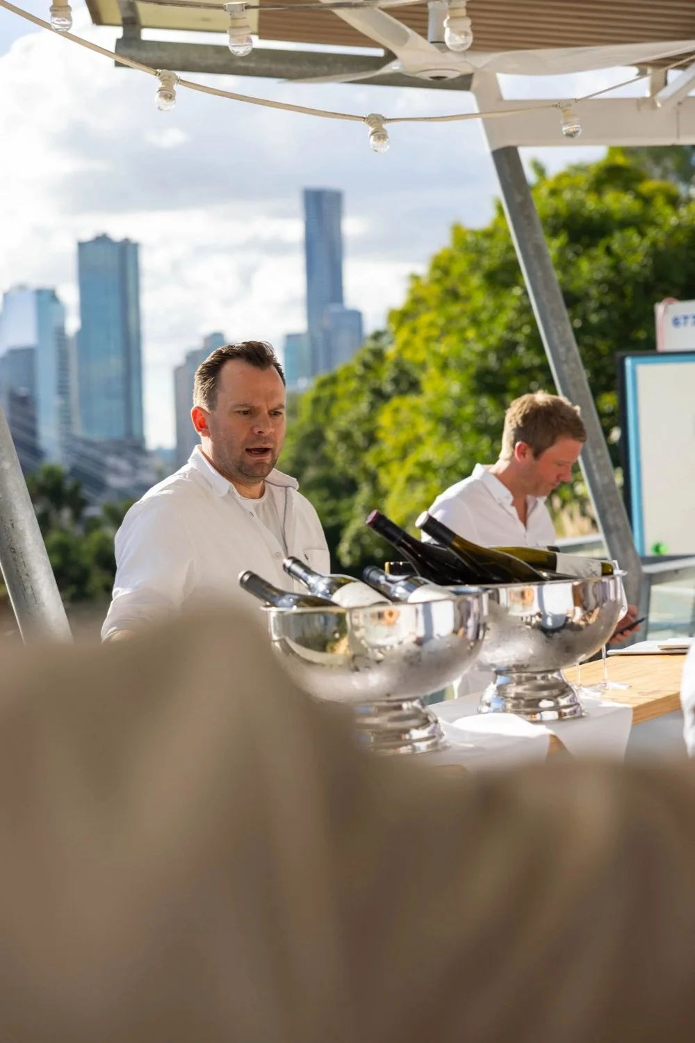 A man with a surprised or confused expression standing at an outdoor bar or wine station, with bottles in silver ice buckets, under a canopy with string lights, and a city skyline with tall buildings and green trees in the background.