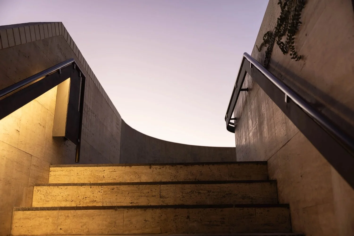 Low-angle view of a modern staircase made of stone, with black metal railings, against a backdrop of a partly lit sky at dusk or dawn.