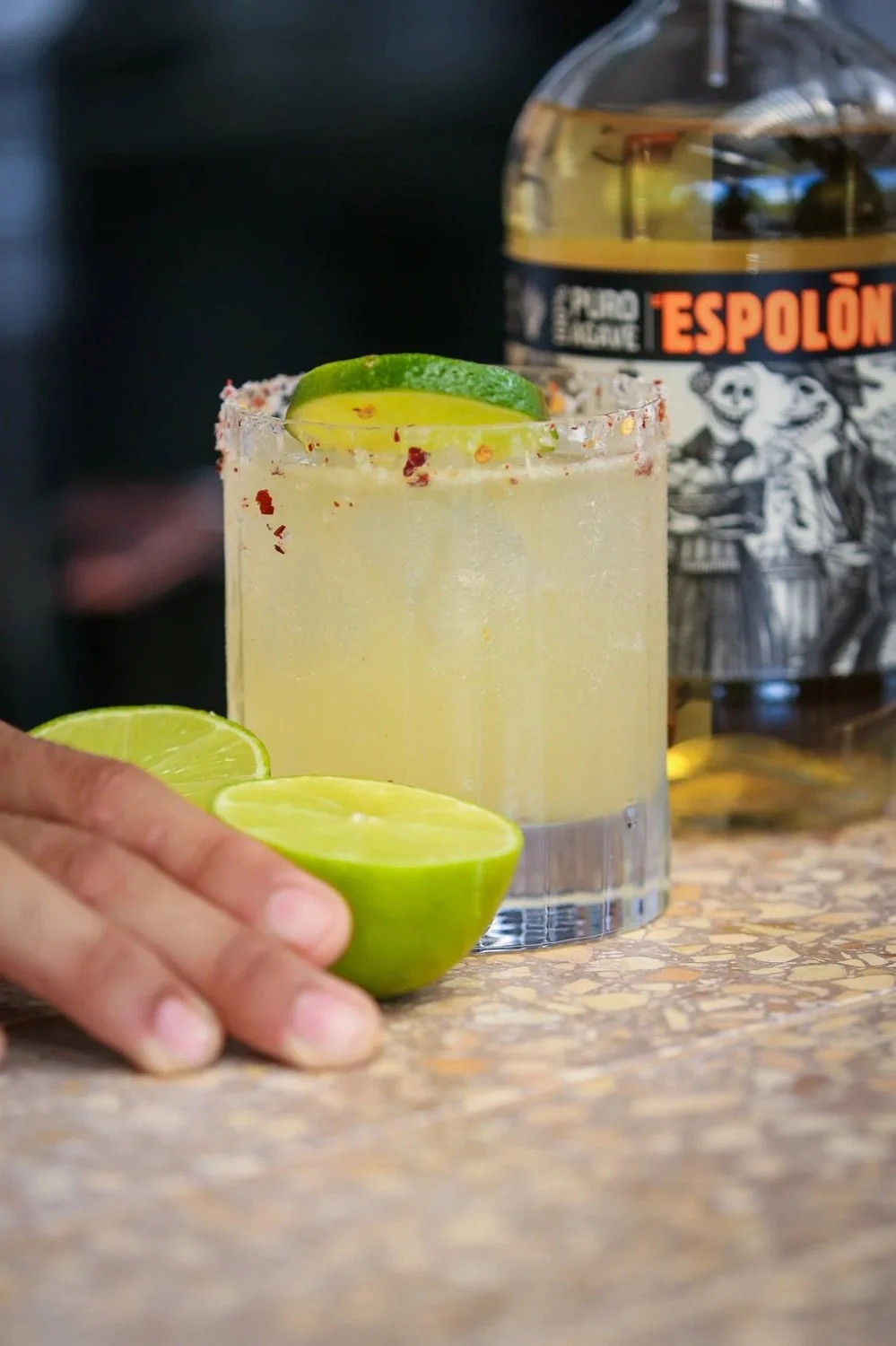 A salted rim glass filled with a yellow citrus beverage, garnished with lime slices and slivers of lime inside, on a speckled countertop with a hand reaching for half a lime. A bottle of Espolòn tequila is in the background.