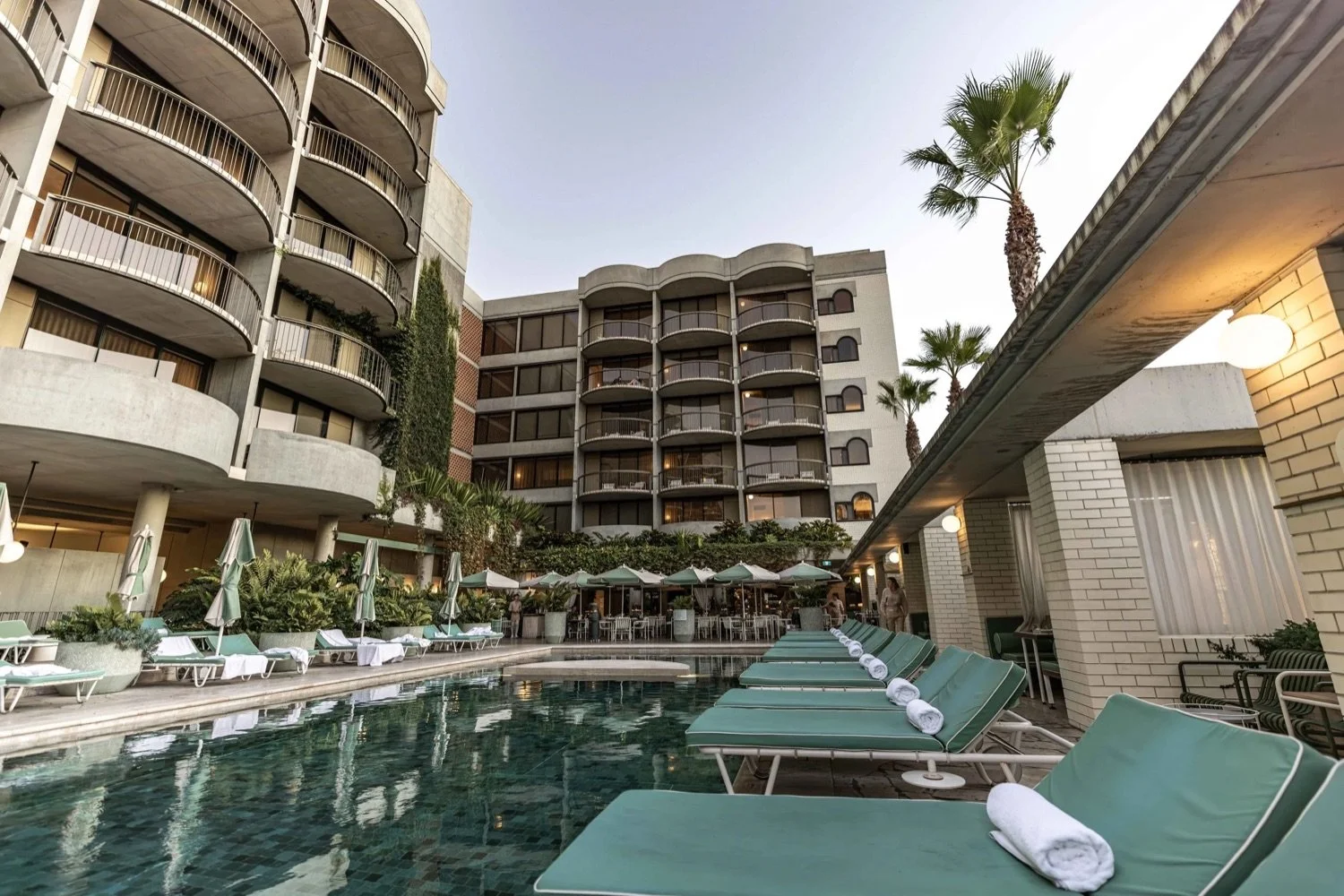 An outdoor hotel pool area with lounge chairs, umbrellas, palm trees, and a multi-story building with balconies.