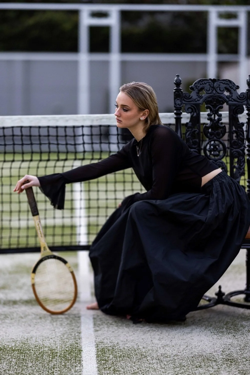 A woman dressed in black, sitting on a tennis court next to a net, holding a vintage tennis racket, with a solemn expression.