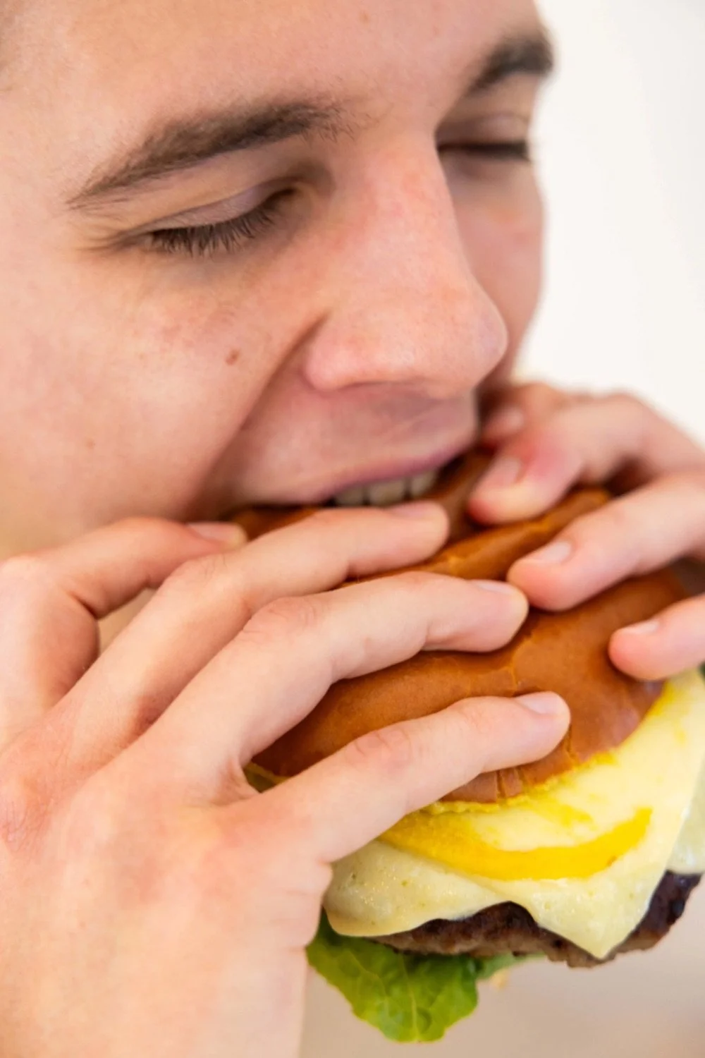 Close-up of a young person with closed eyes biting into a large cheeseburger with lettuce, cheese, burger patty, egg, and bun.