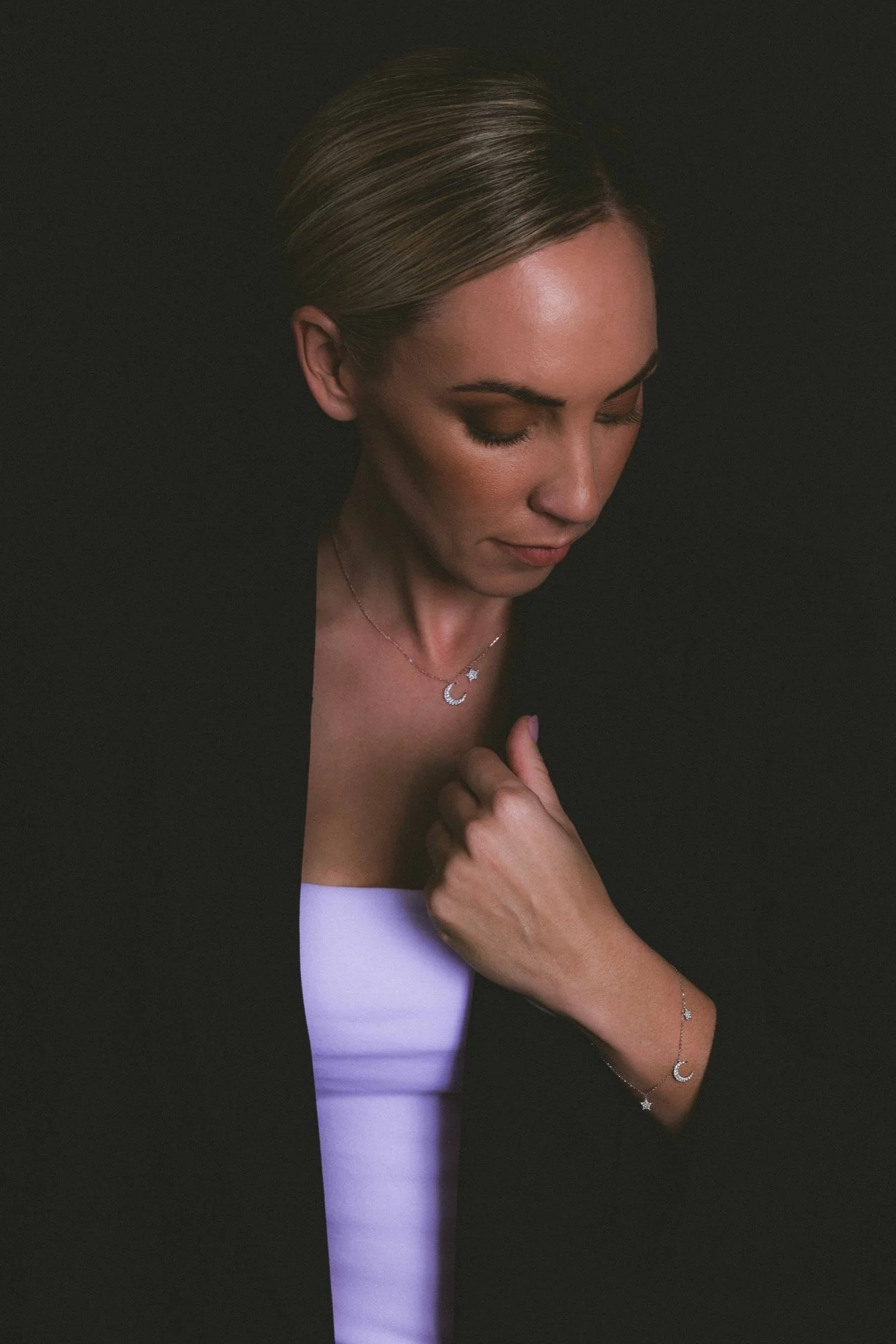 A woman with blonde hair styled in an updo, wearing a black blazer over a white top, showcasing moon and star jewelry including a necklace, bracelet, and earrings against a dark background.