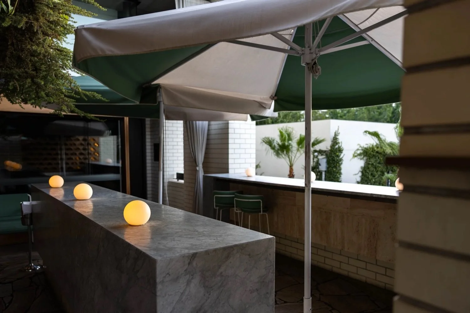 An outdoor patio with a marble bar counter illuminated by glowing spherical lights, green umbrellas providing shade, and a background of white brick walls, green plants, and outdoor furniture.