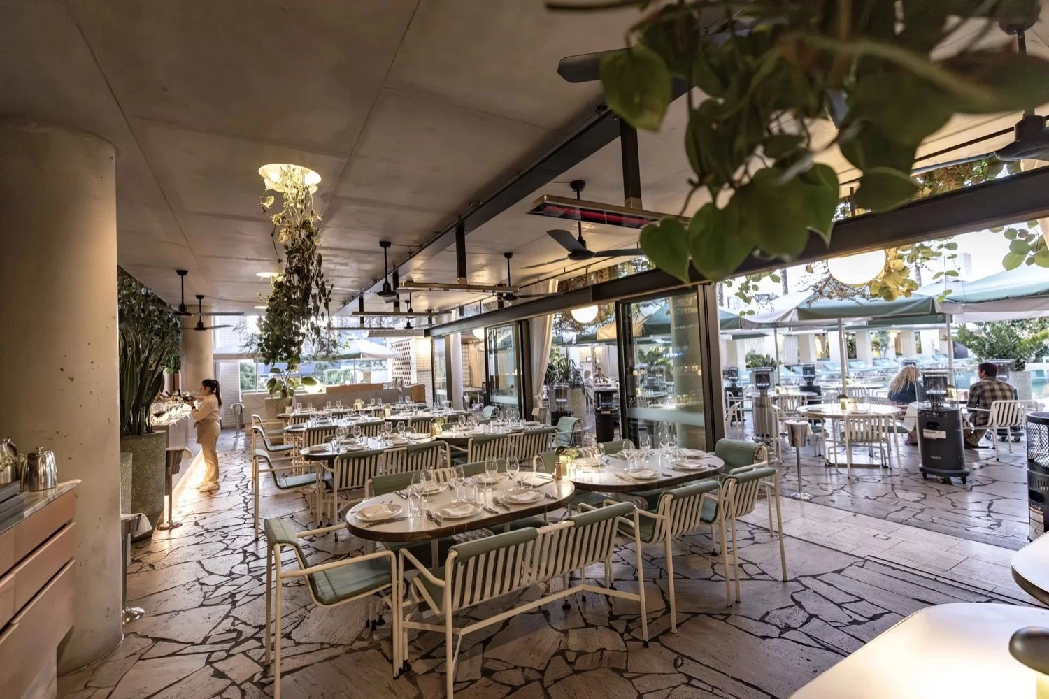 Interior of a stylish restaurant with tables set for dining, large windows, and an outdoor patio area visible through the windows with tables, chairs, and umbrellas.