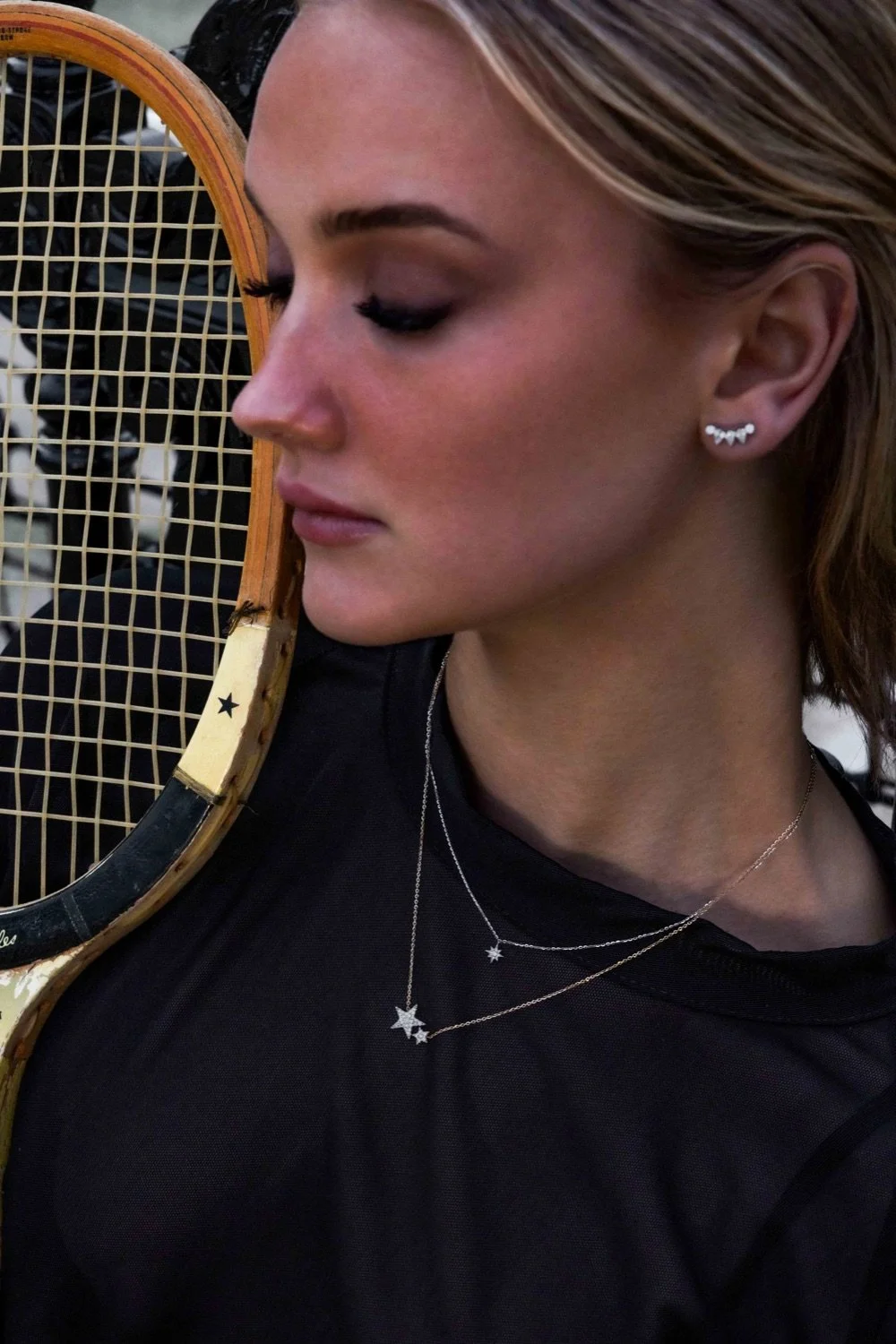 A woman with light makeup and earrings, holding a tennis racket near her face.