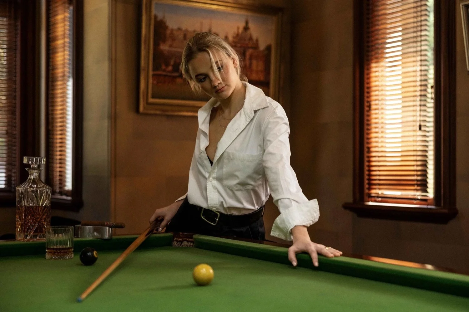 A woman in a white shirt playing pool in a dimly lit room with wooden decorations and window blinds.