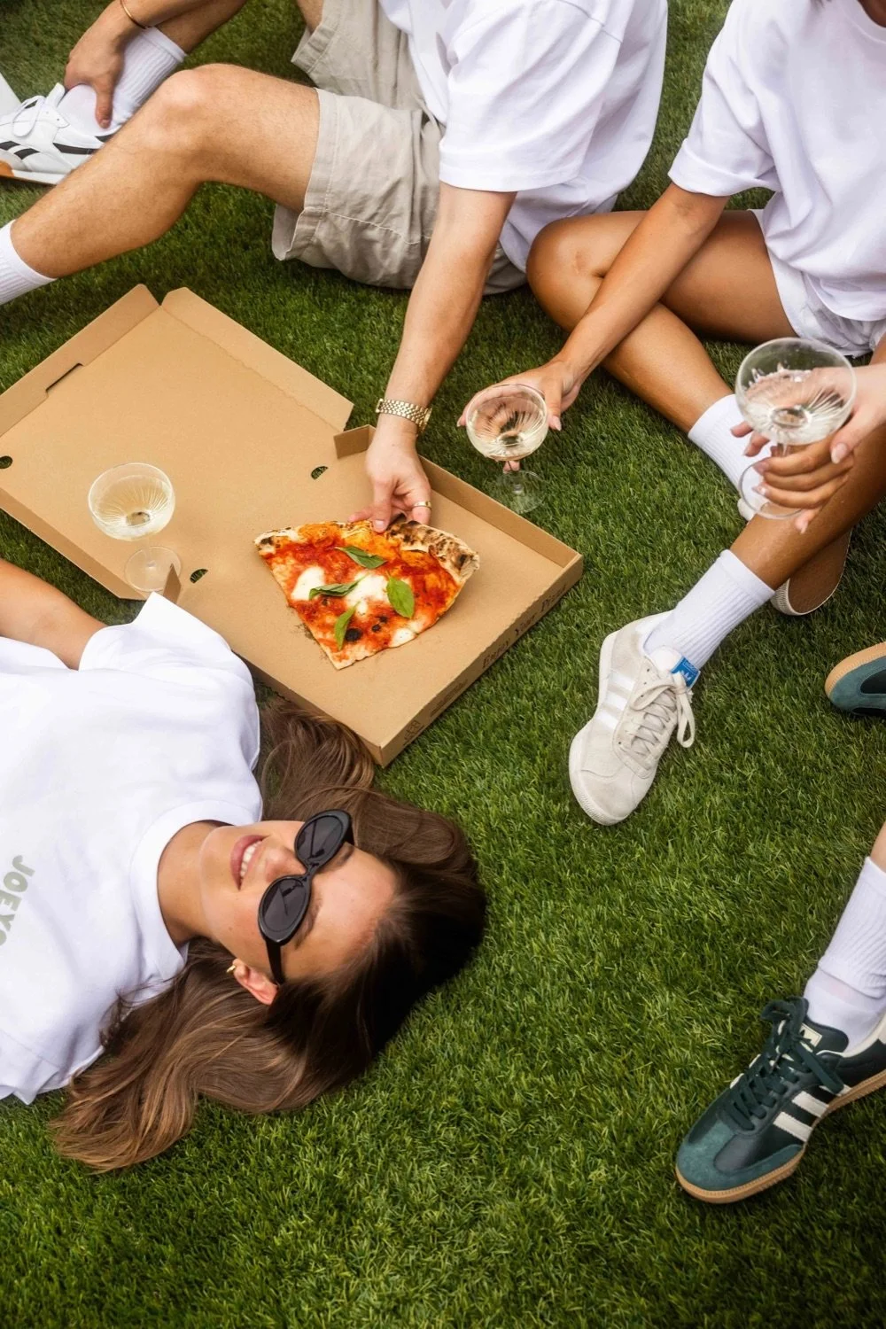 A woman lying on the grass with sunglasses, smiling at a picnic table. Nearby, a group of people in white shirts and shorts are sitting on the grass, reaching for a slice of pizza from an open pizza box, with glasses of white wine around.