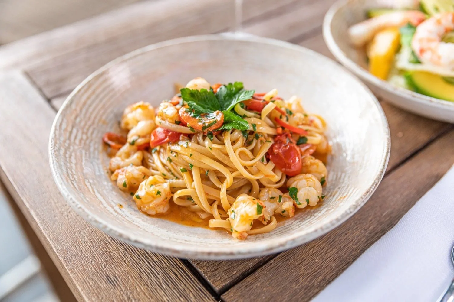 A bowl of shrimp pasta garnished with parsley and cherry tomatoes on a wooden table with another bowl of salad in the background.