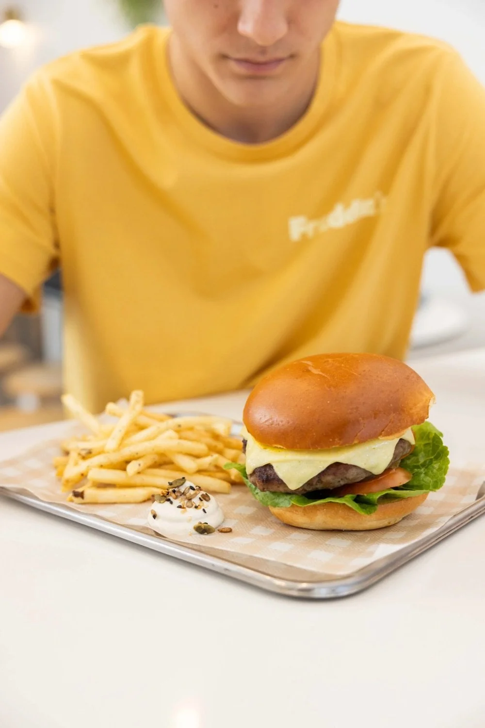 A hamburger with a beef patty, cheese, lettuce, tomato, and a sandwich bun, served with French fries and a small dollop of sauce topped with seeds on a tray.
