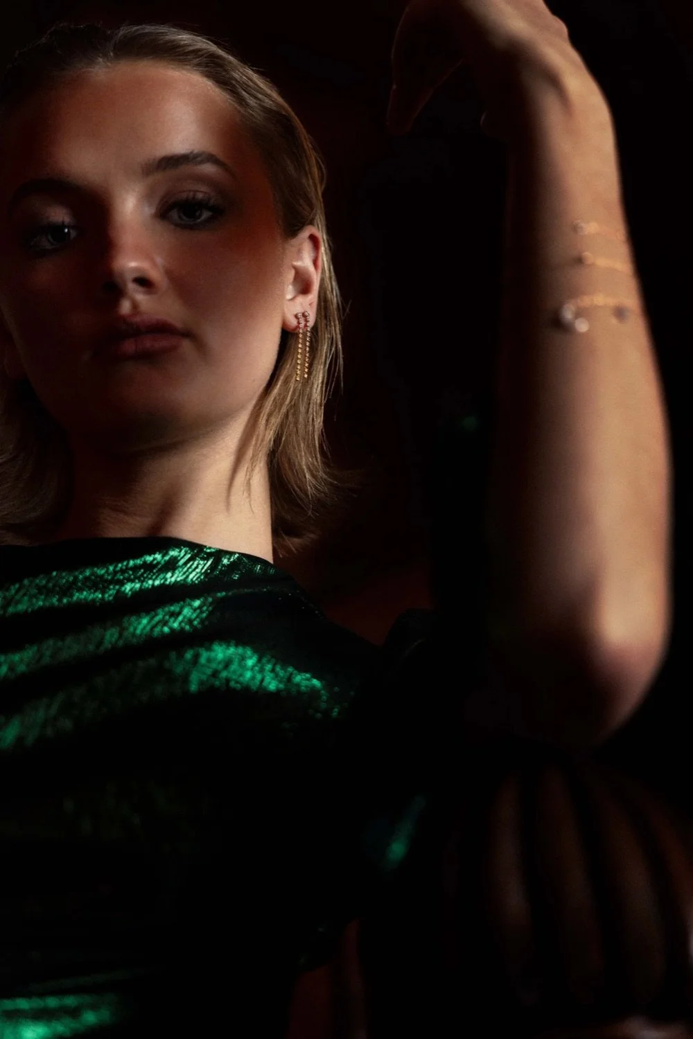 Close-up of a young woman with short blond hair, wearing gold earrings and a shiny black and green top, posing with her left arm raised against a dark background.