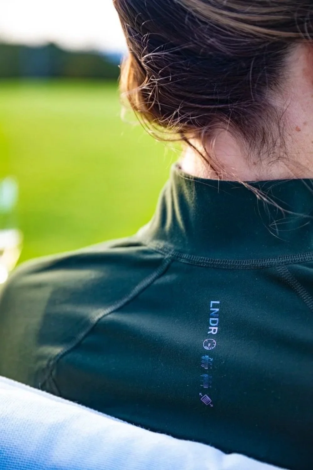 Close-up of a person wearing a black jacket with the brand logo 'Lndr' and Scottish weather icons, sitting outdoors on a sunny day with a green field in the background.
