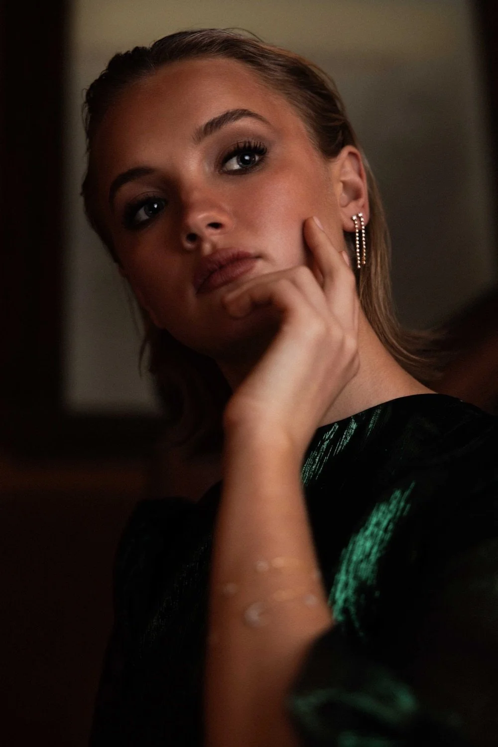 A young woman with light skin and blonde hair, wearing earrings and a dark-colored top, appears to be deep in thought, resting her chin on her hand.