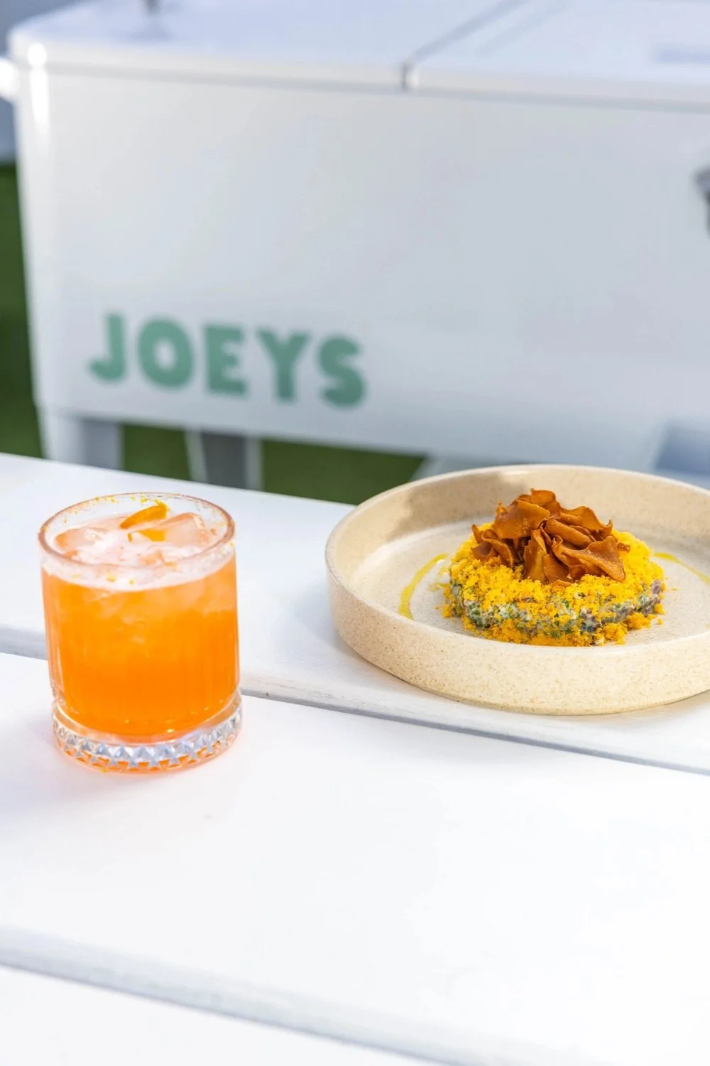 A vibrant orange cocktail with ice and a lemon twist in a textured glass, placed next to a plate with a colorful salad topped with shredded meat and yellow crumble, on a white table outdoors, with a blurred white sign in the background reading 'JOEYS