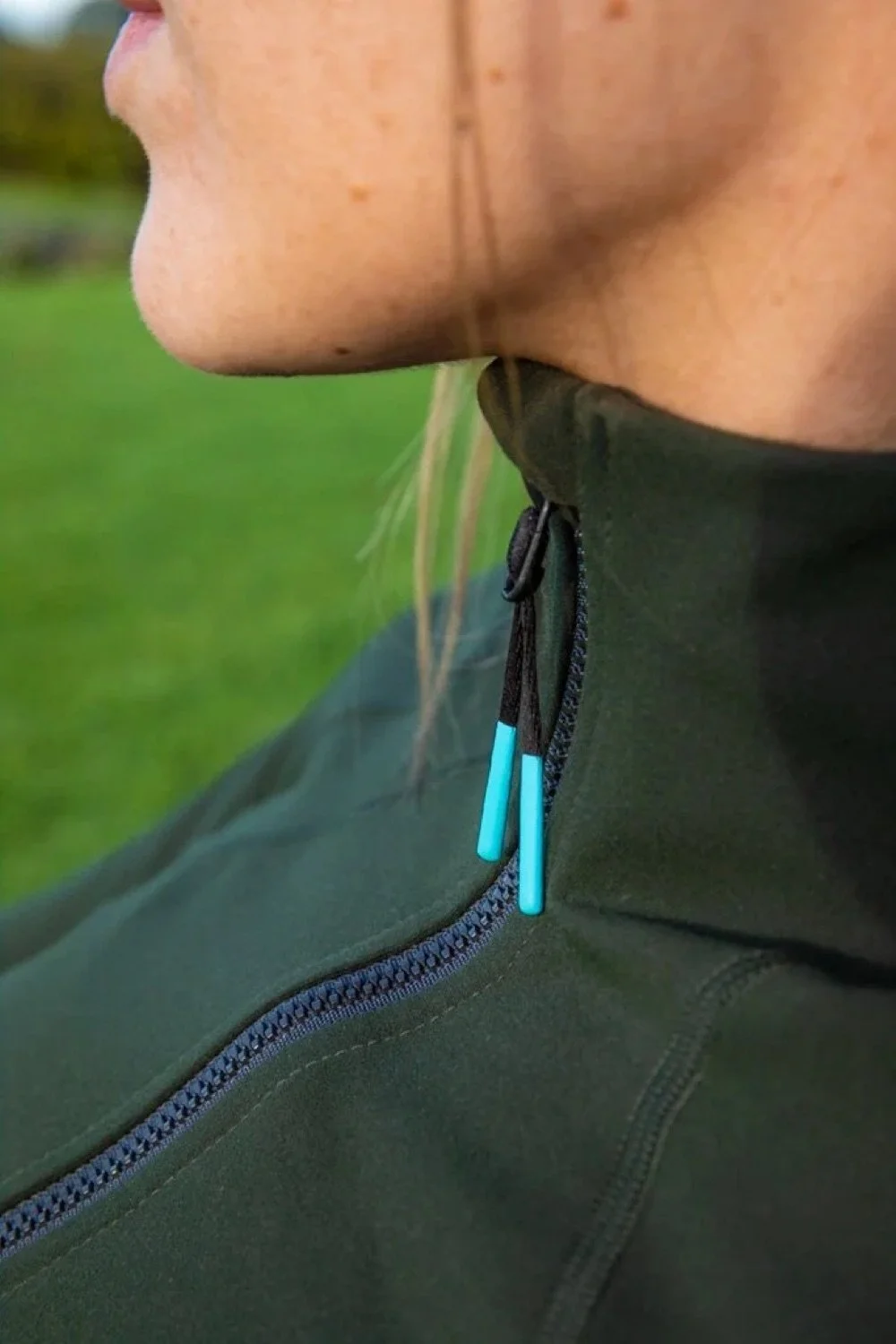 Close-up of a person's neck and chin wearing a green outdoor jacket with a zipper and blue zipper pulls, with a blurred green outdoor background.