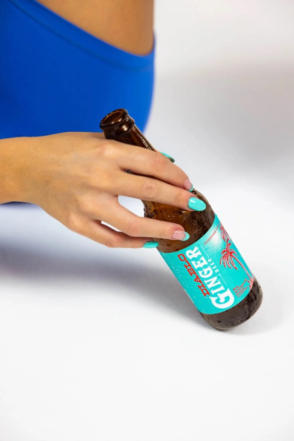 A person with turquoise-painted nails holding a brown glass bottle of Ginger beer labeled "DABBIO" on a white surface.
