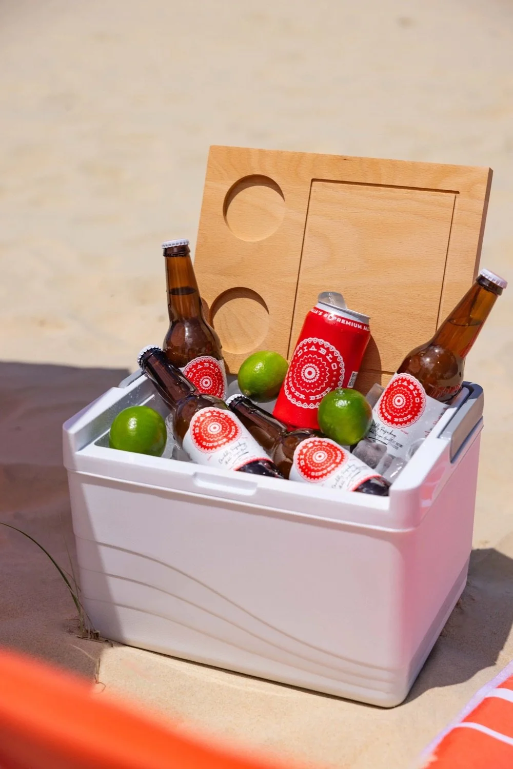 A white cooler filled with beer bottles, a red can, limes, and a wooden cutting board, placed on sand.