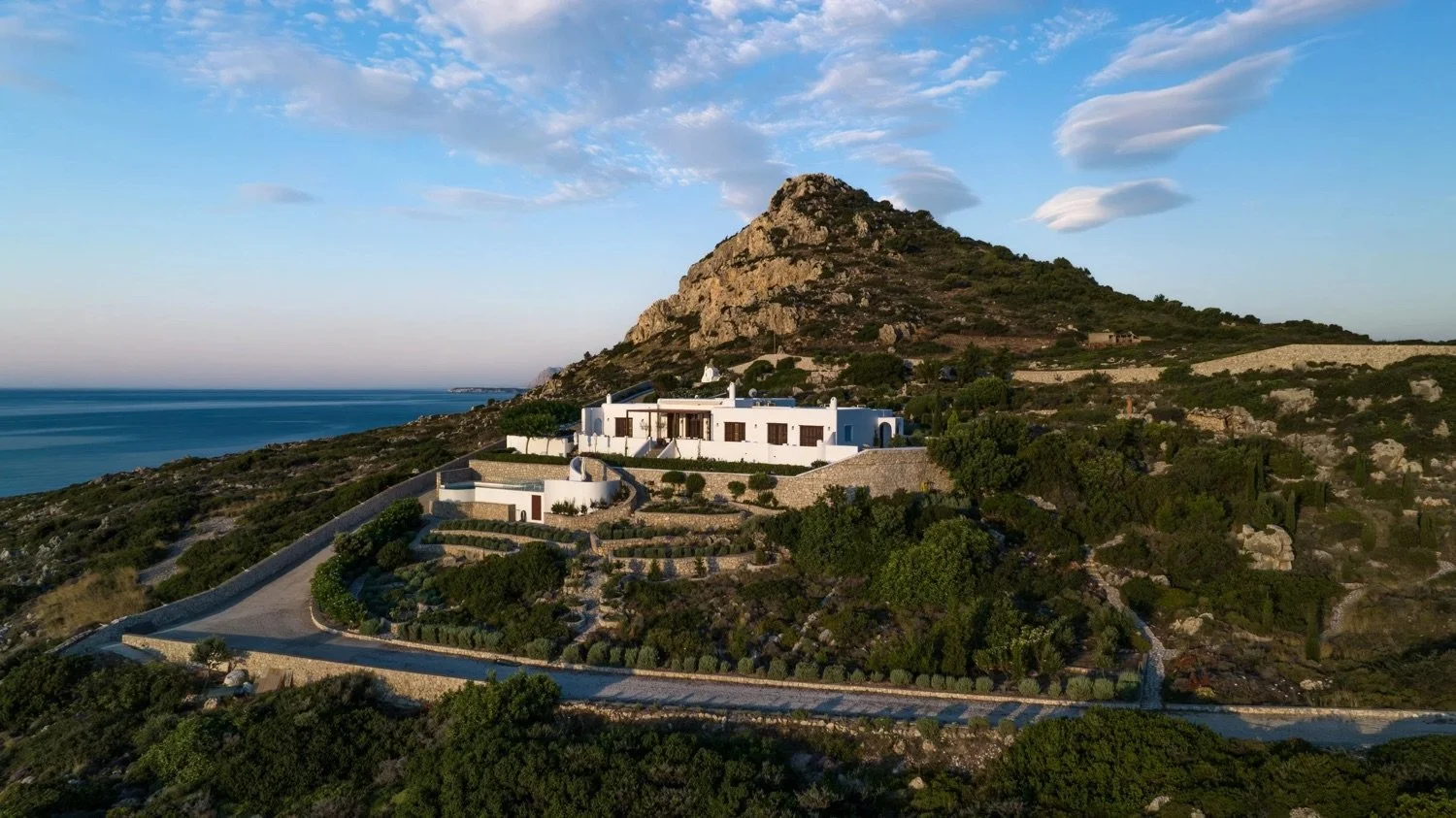 A white house situated on a hillside with a winding driveway, surrounded by greenery, with a mountain and the ocean in the background during sunset.