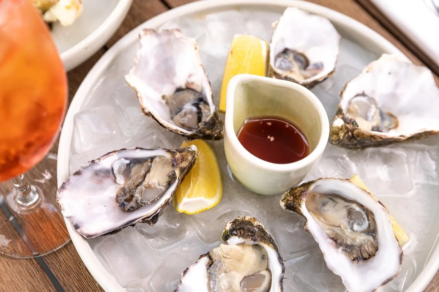 A platter of raw oysters on ice with lemon wedges and a small pitcher of cocktail sauce in the center.