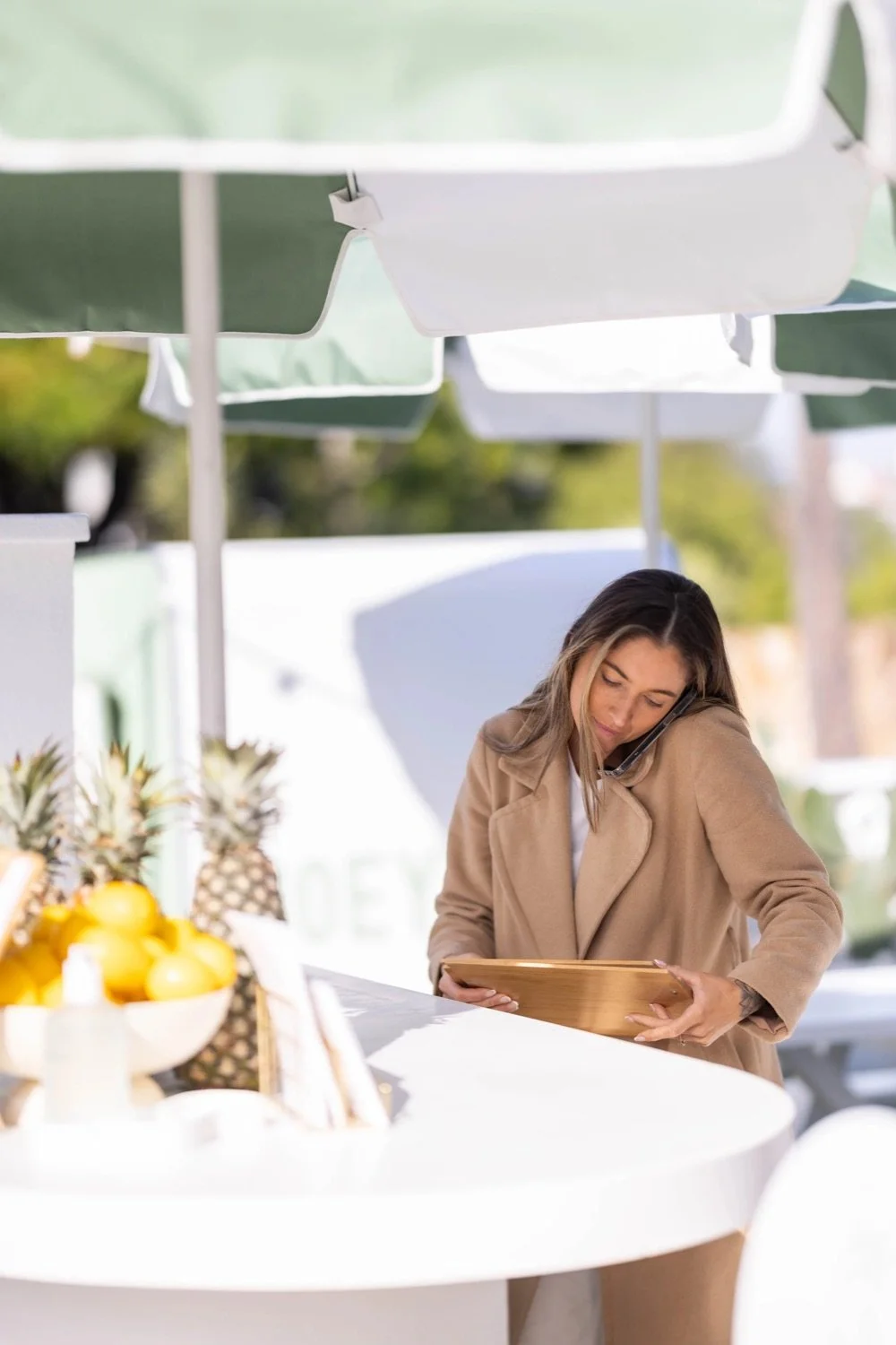 Woman in beige coat talking on a cell phone while looking at a wooden tray at an outdoor market stand with fruit and pineapples.