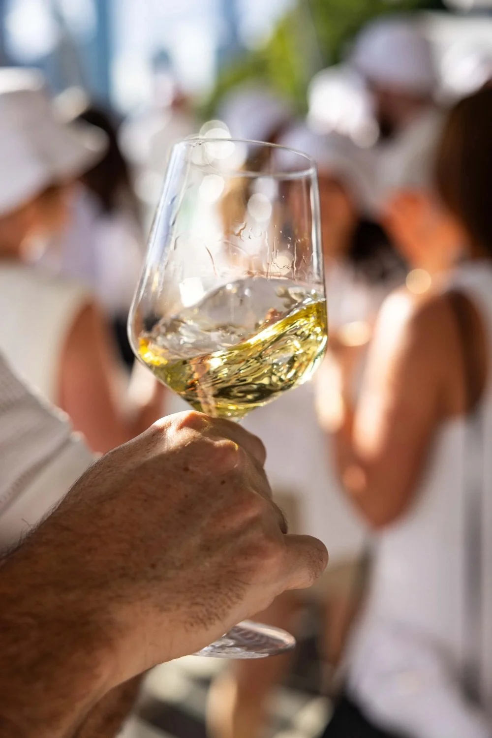 A person's hand holding a glass of white wine, with a blurred group of people in the background at a social gathering.