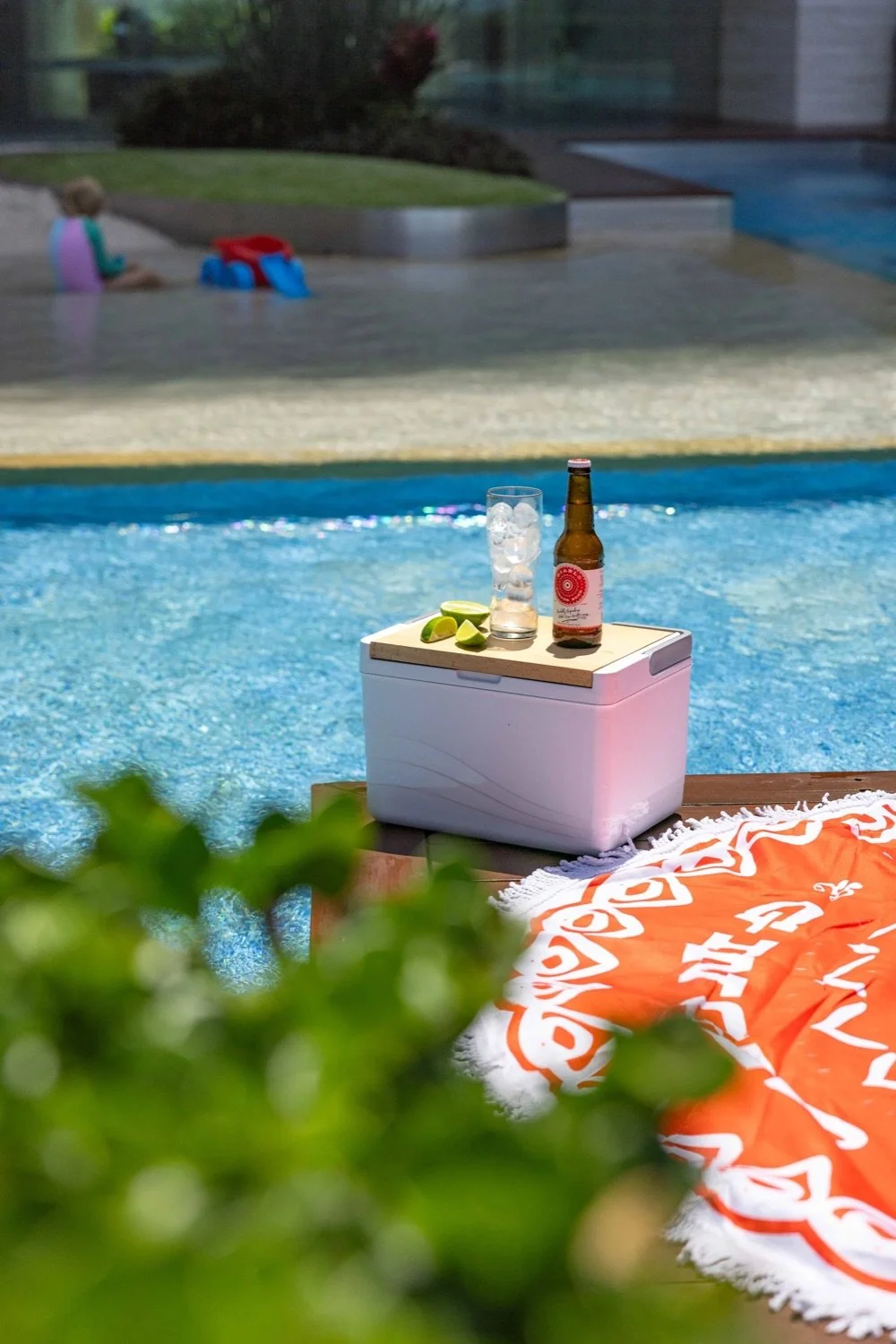 A poolside scene with a small white cooler holding a bottle of beer, a glass of ice and soda, and lime wedges on top. An orange and white towel with beach designs is partially visible on the right. In the background, a small child sits on the pool de