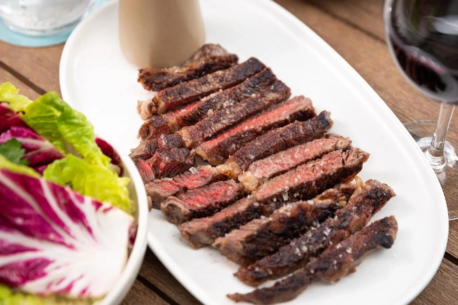 Sliced medium-rare steak on a white platter with a side salad and glass of red wine.