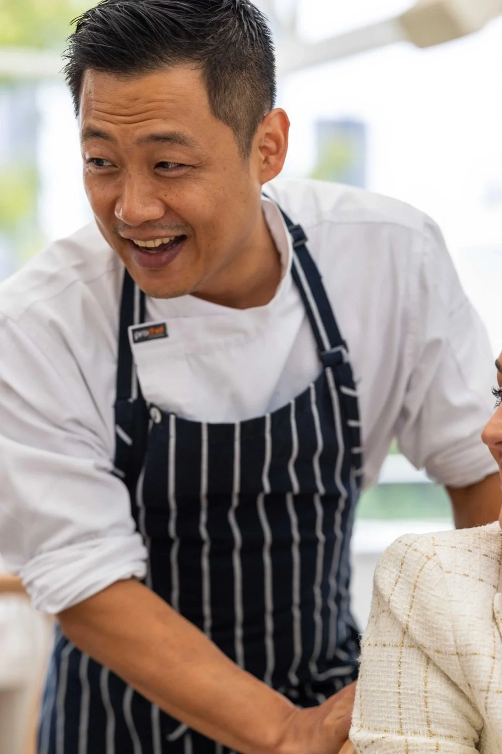 A man wearing a striped apron and white shirt leaning over a seated woman, smiling and engaging in conversation, in a bright, modern indoor setting.