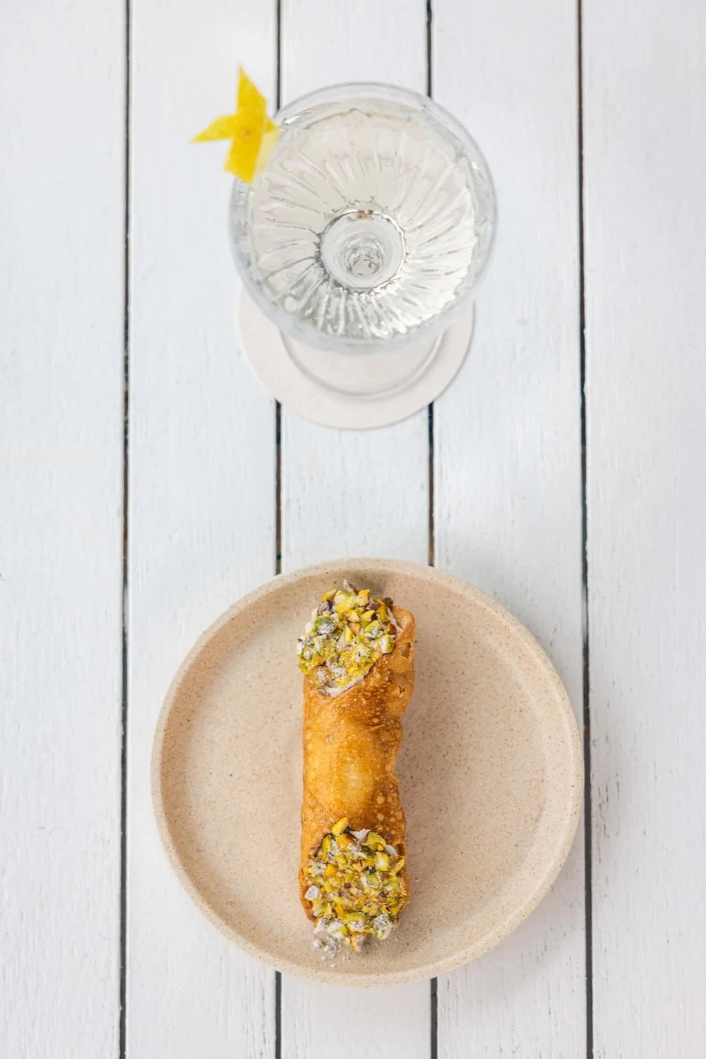 A glass of water with a lemon wedge and a plated dessert topped with chopped pistachios on a white wooden surface.