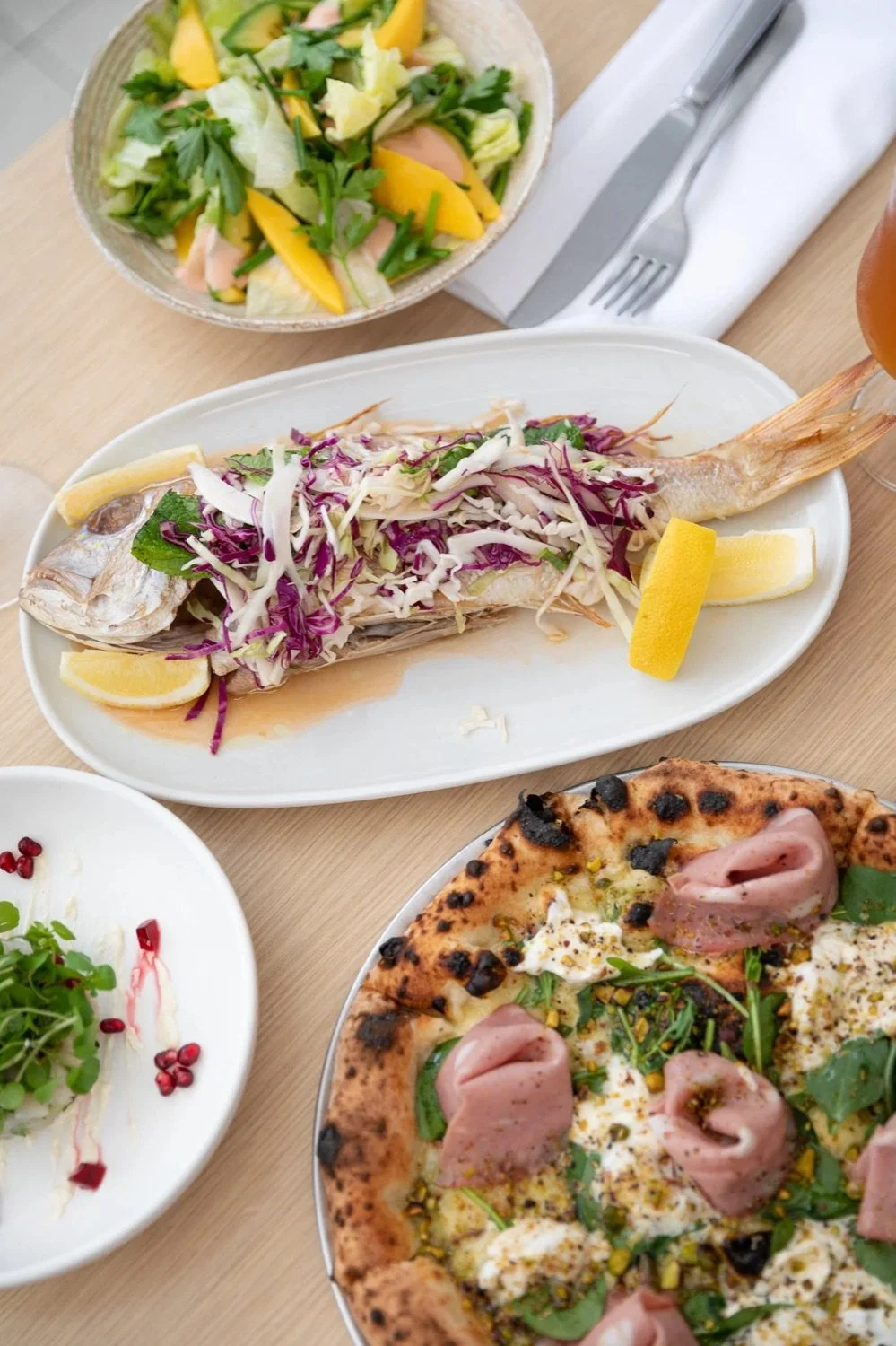 A table with various dishes including a whole grilled fish topped with shredded cabbage and lemon wedges, a fresh salad with mixed greens and mango slices, a pizza with ham and greens, and a small salad with microgreens and pomegranate seeds.