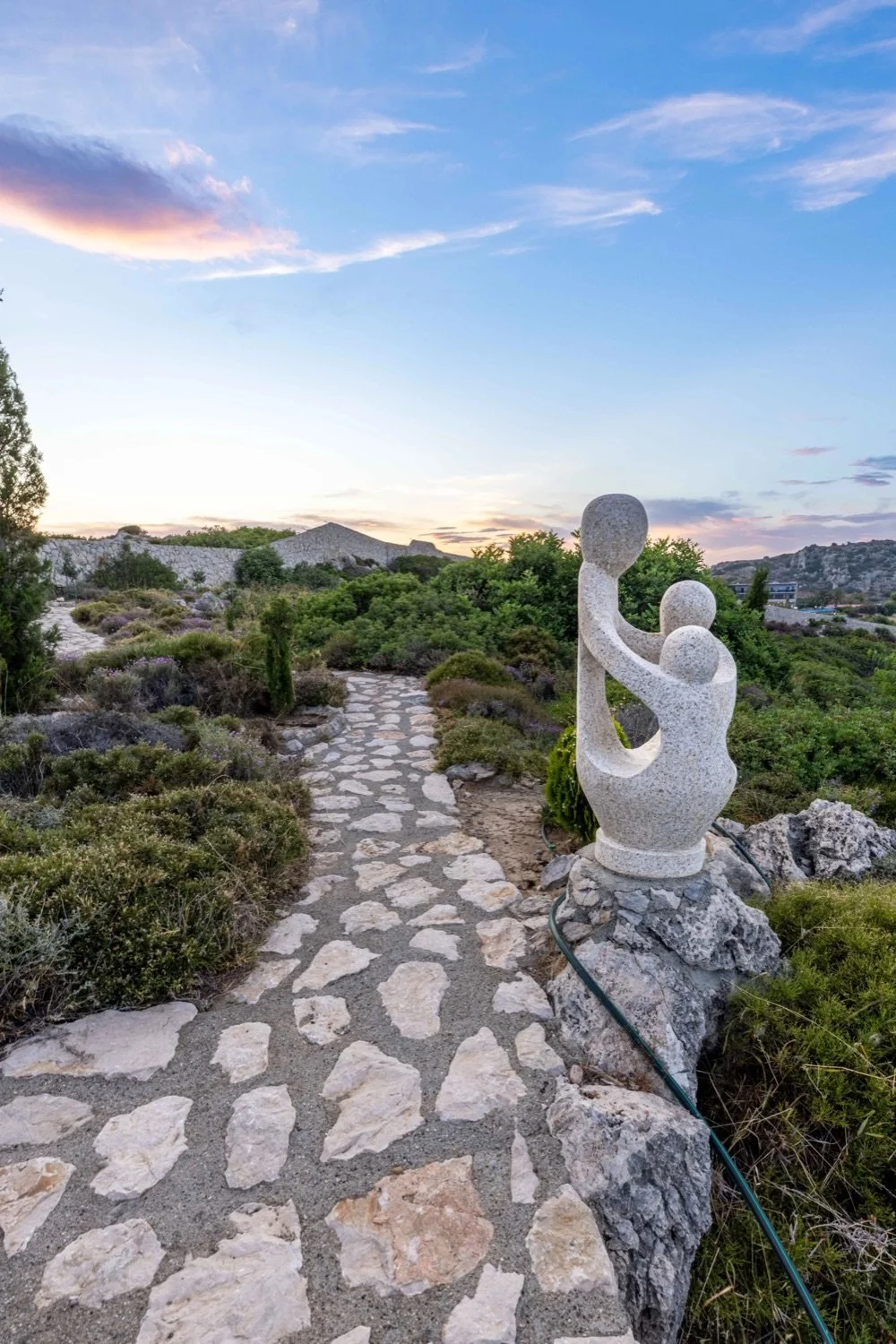 A stone sculpture resembling abstract human figures holding hands, located on a rocky pathway surrounded by desert vegetation under a colorful sky at sunset.
