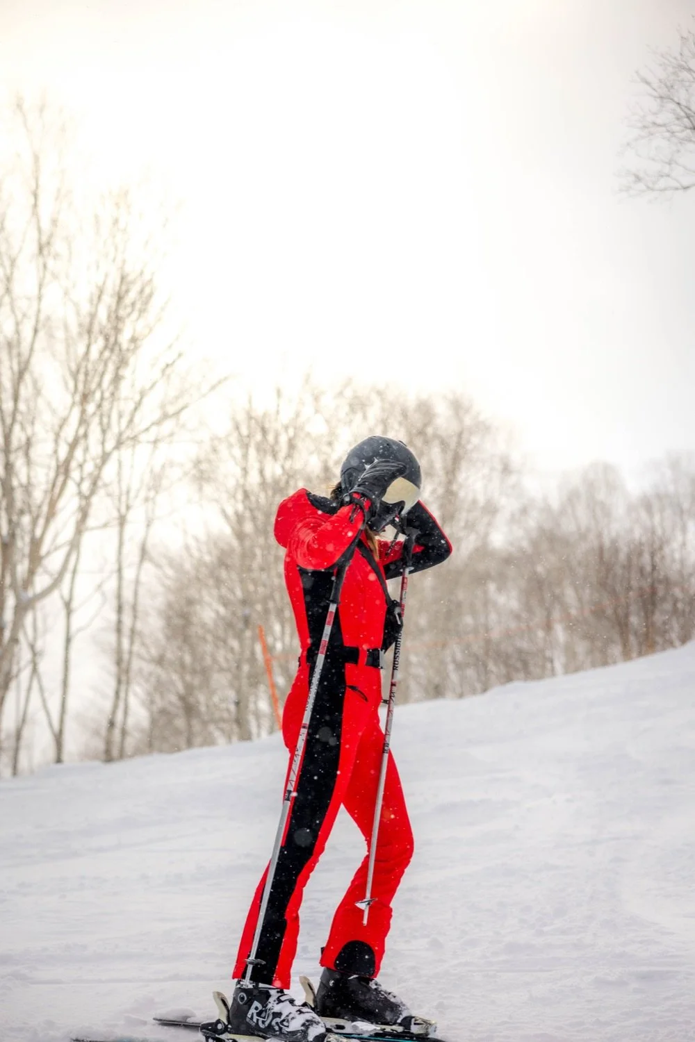 A person dressed in a red ski suit and black helmet, standing on skis in a snowy landscape, adjusting their helmet with snow falling around.