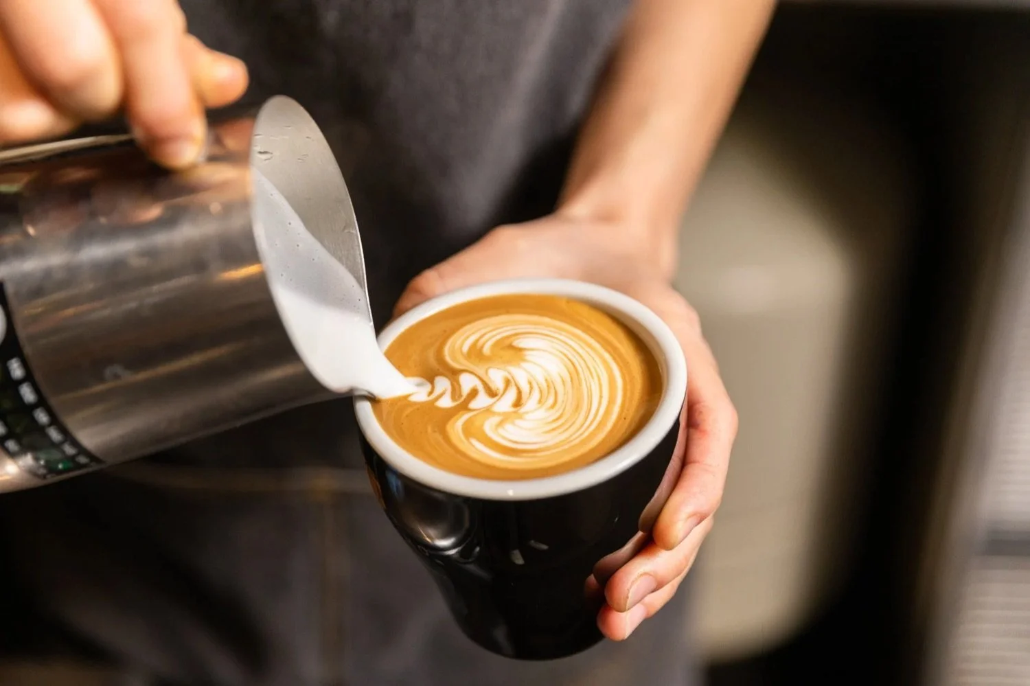 Person pouring steamed milk into a cup of coffee, creating latte art with a swirling pattern.