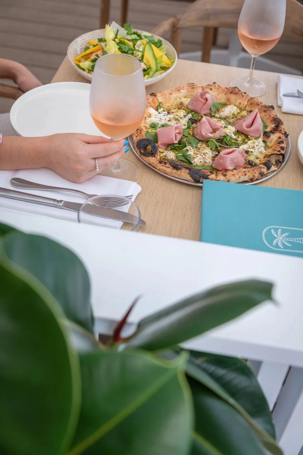 A table set with a pizza topped with ham and greens, two glasses of rosé wine, and a bowl of salad with lettuce, mango, and cucumber. A person's hand with a ring is holding one glass of wine.