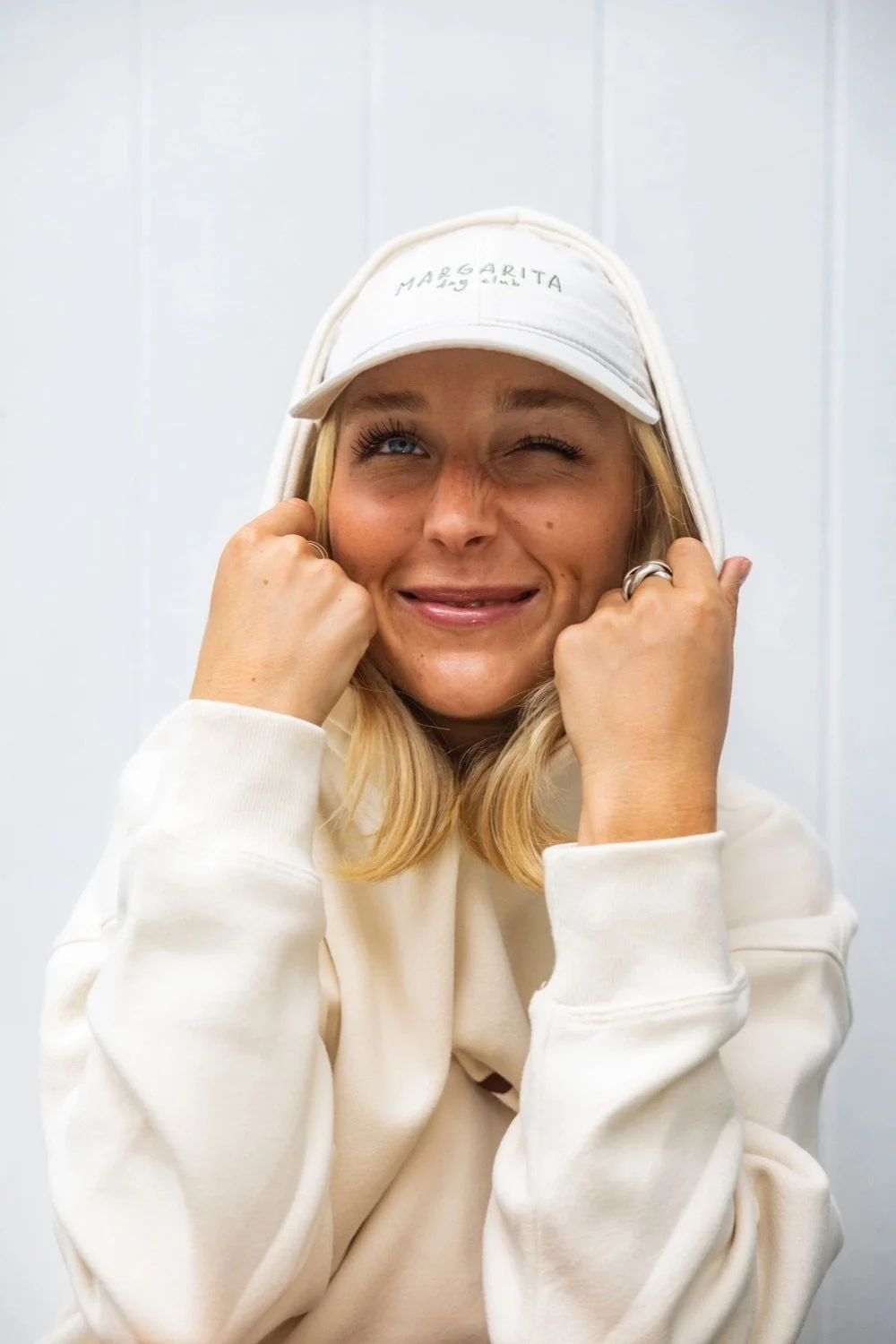 A woman with blonde hair wearing a white hoodie and a white cap that says 'Margarita' making a silly facial expression.