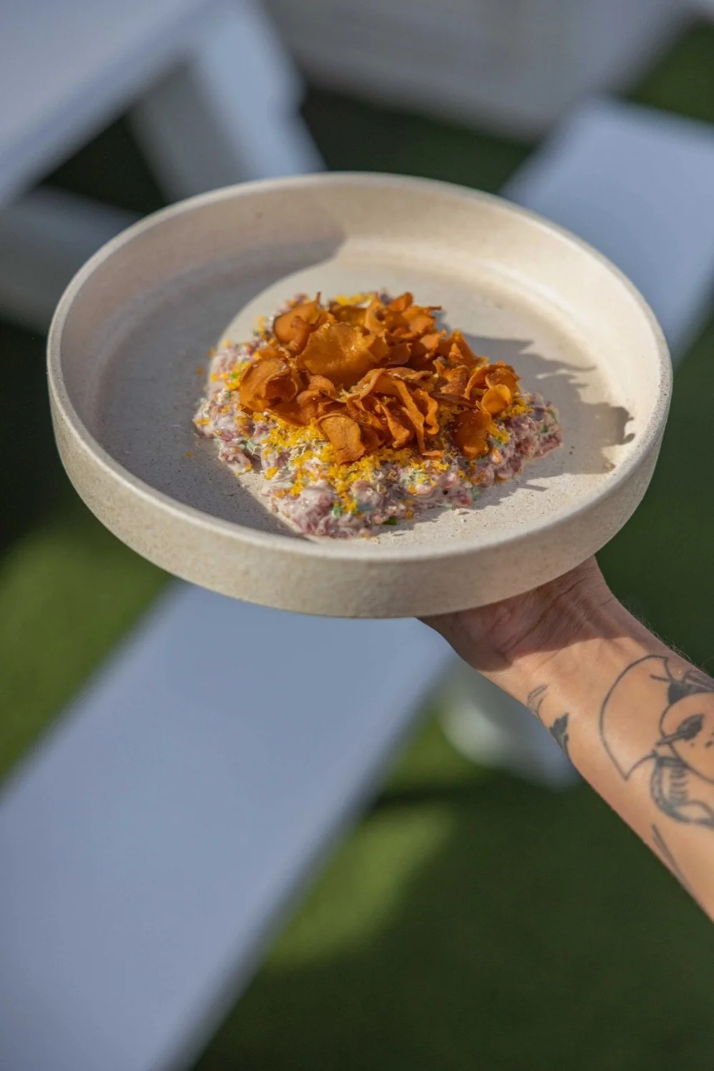 A hand holding a beige bowl with a dessert topped with caramelized slices on an outdoor table.