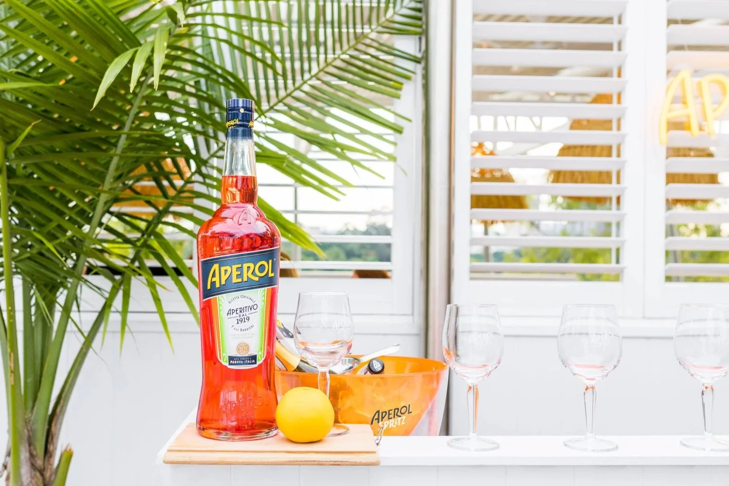 A bottle of Aperol, an orange lemon, four empty glasses, and an orange bowl on a white countertop, with a large green palm leaf and white window shutters in the background.