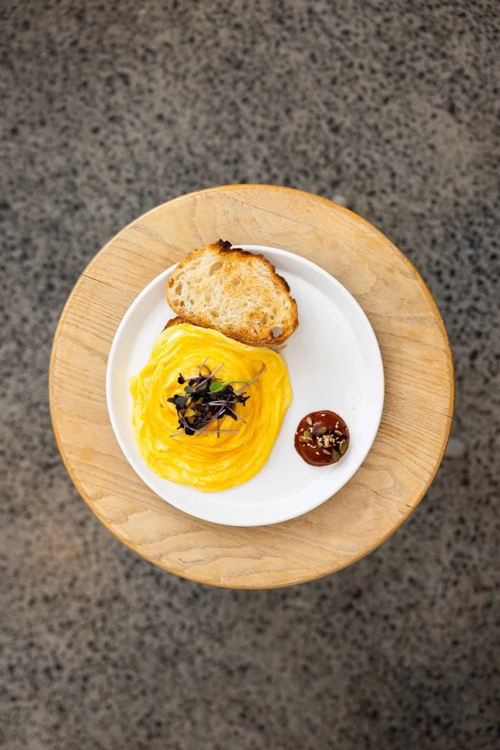 A plate contains a serving of scrambled eggs garnished with microgreens, a slice of toasted bread, and a small dollop of sauce topped with seeds, on a round wooden table.