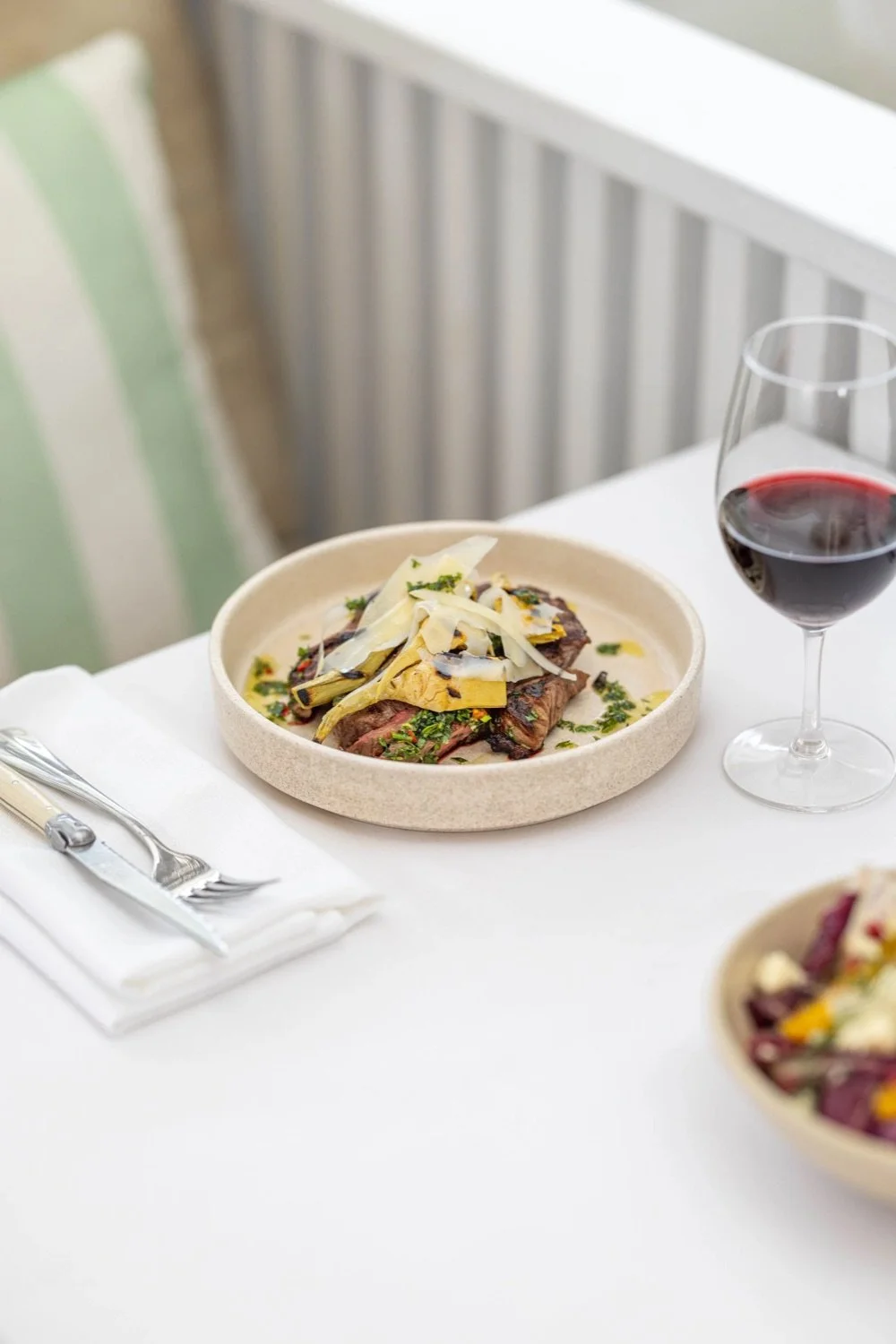 A plate of beef steak topped with cheese, herbs, and vegetables on a white tablecloth next to a glass of red wine.