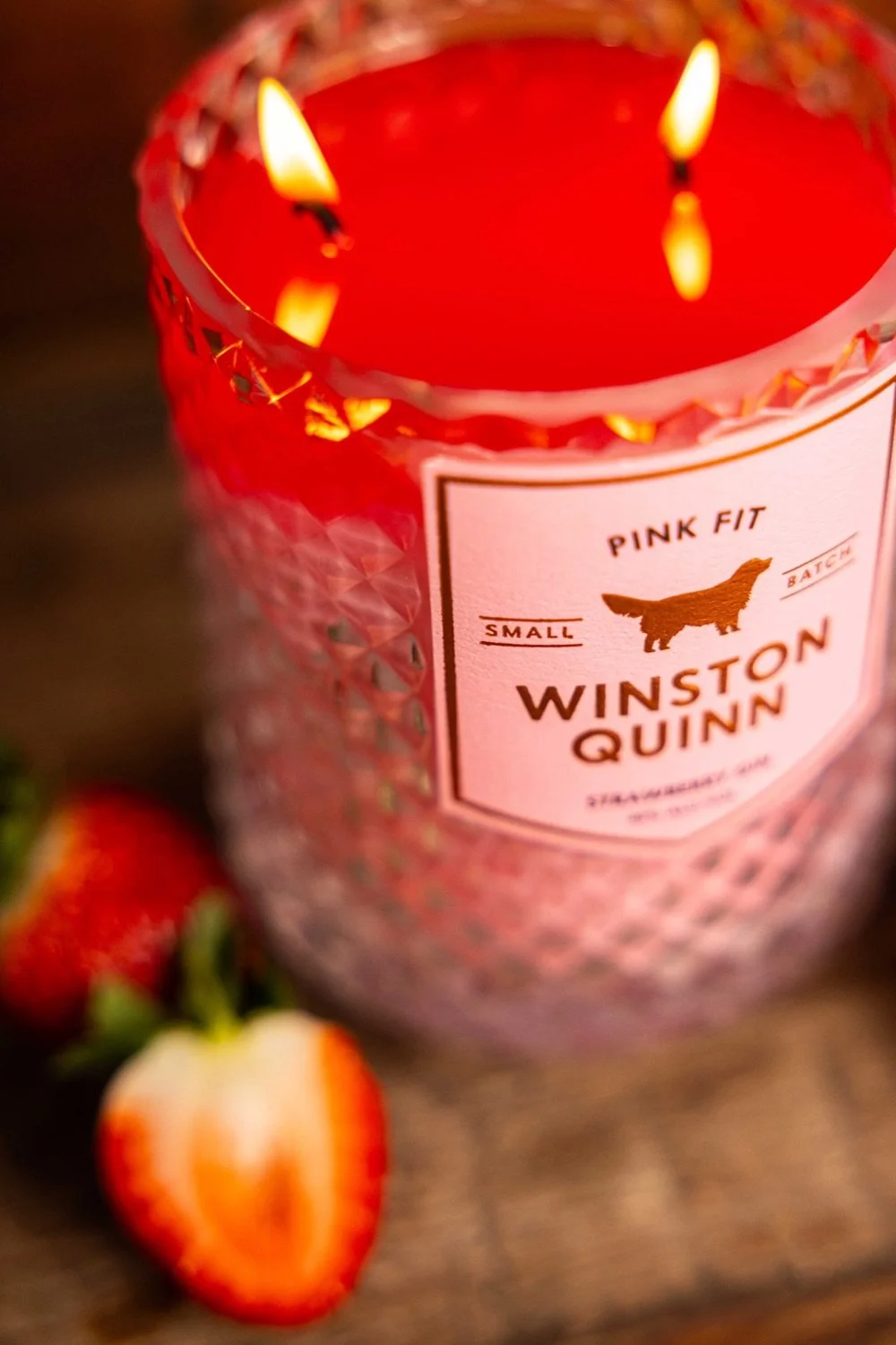 A lit pink candle with a textured glass holder, labeled 'Winston Quinn' and 'Pink Fit'. Strawberries are placed nearby on a wooden surface.