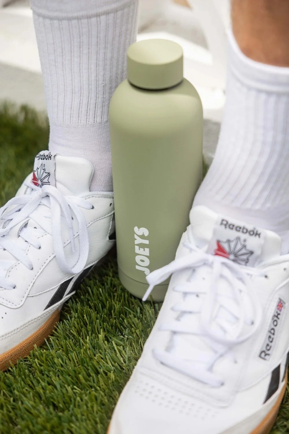 A close-up of white Reebok sneakers, white socks, and a green water bottle with the word 'JOFYS' on it, all on green grass.
