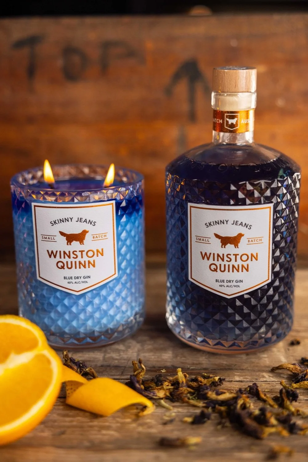A lit blue glass candle and a blue bottle of Winston Quinn Blue Dry Gin on a rustic wooden surface, with lemon slices and dried tea leaves nearby, and a wooden background with an arrow pointing up and the word 'TOP' written.