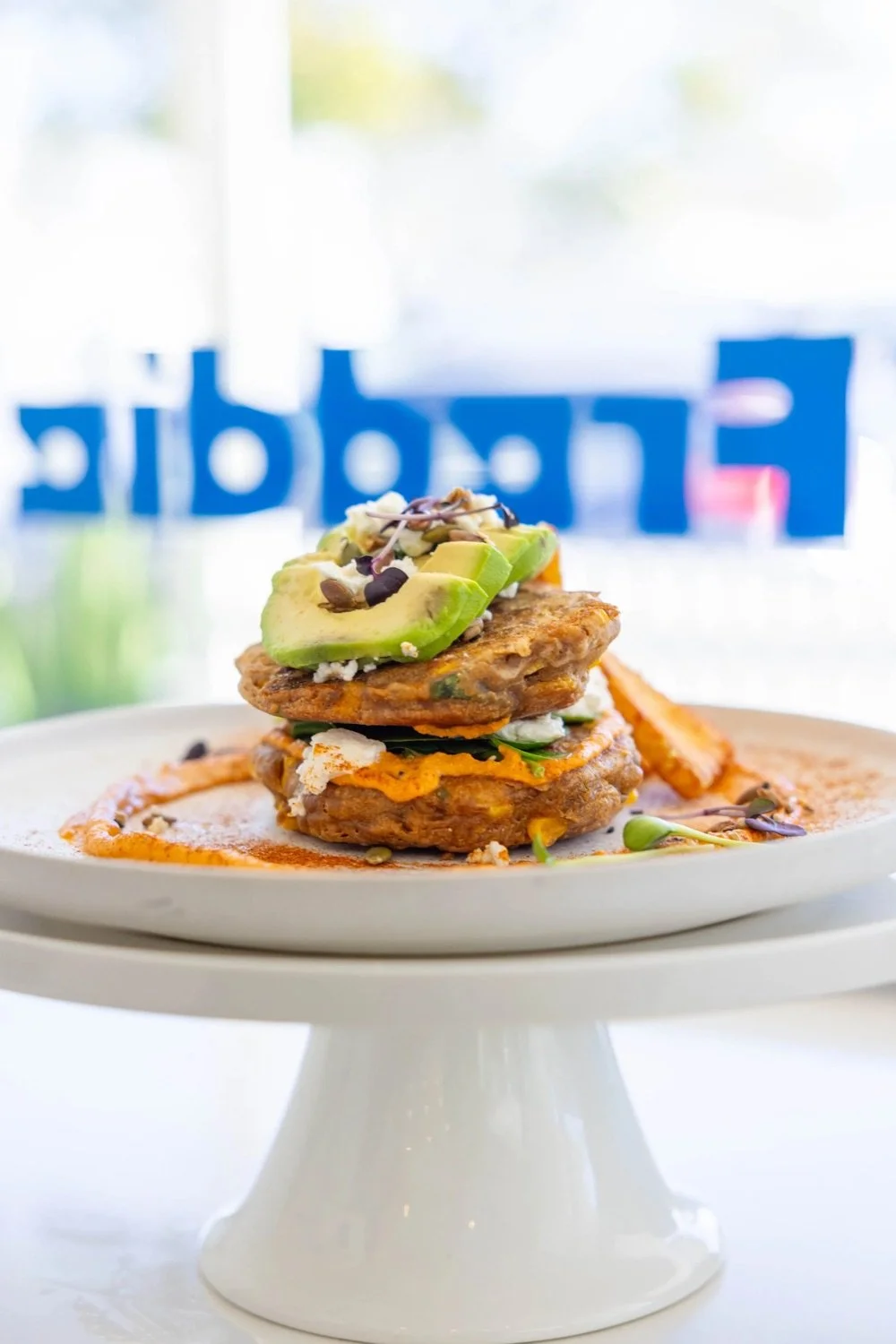 A plate of stacked veggie burger patties topped with sliced avocado, microgreens, and drizzled with sauce, set on a white cake stand.