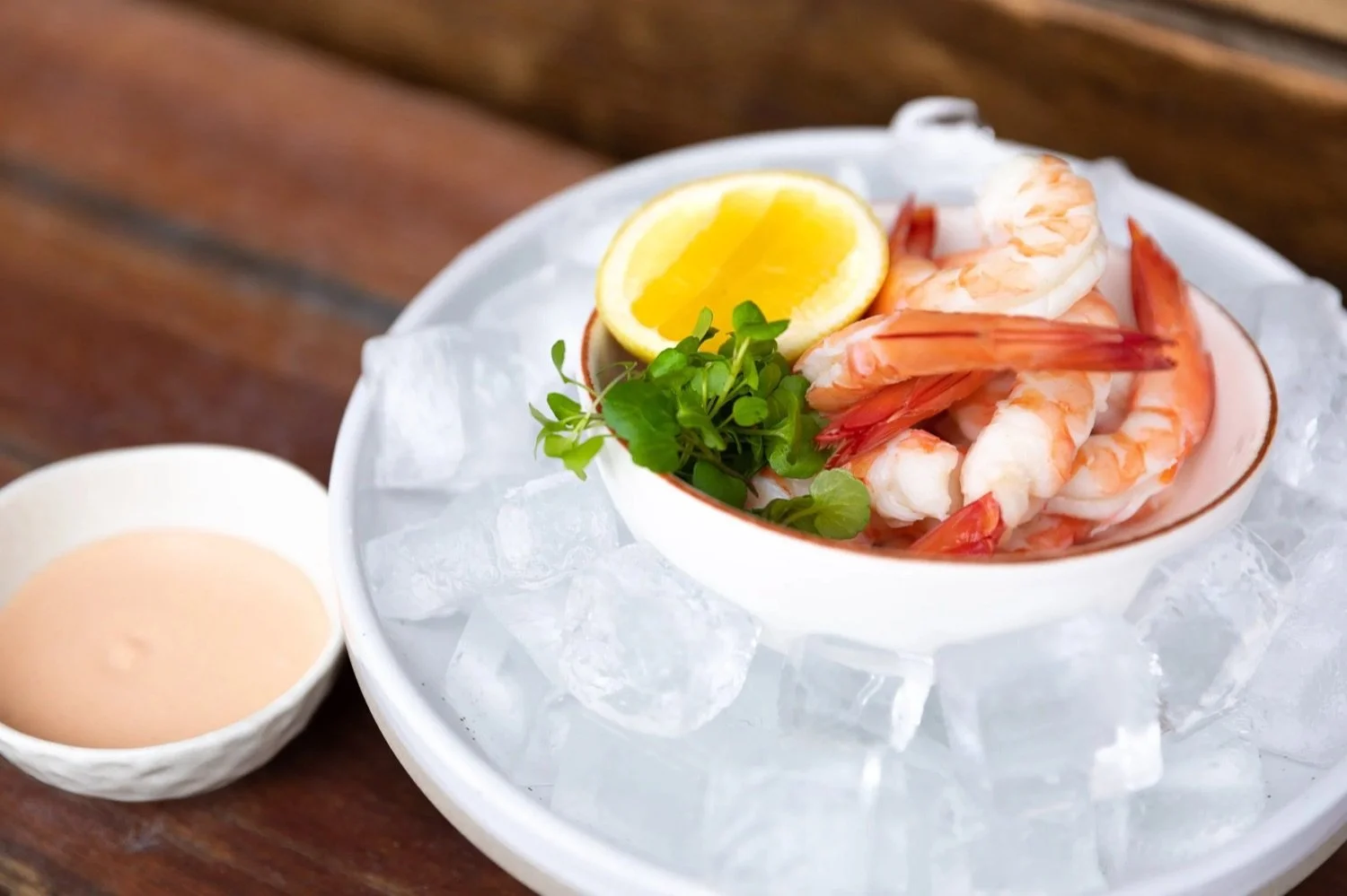 A bowl of cooked shrimp garnished with lemon wedge and herbs, resting on a bed of ice with a small dish of dipping sauce beside it.