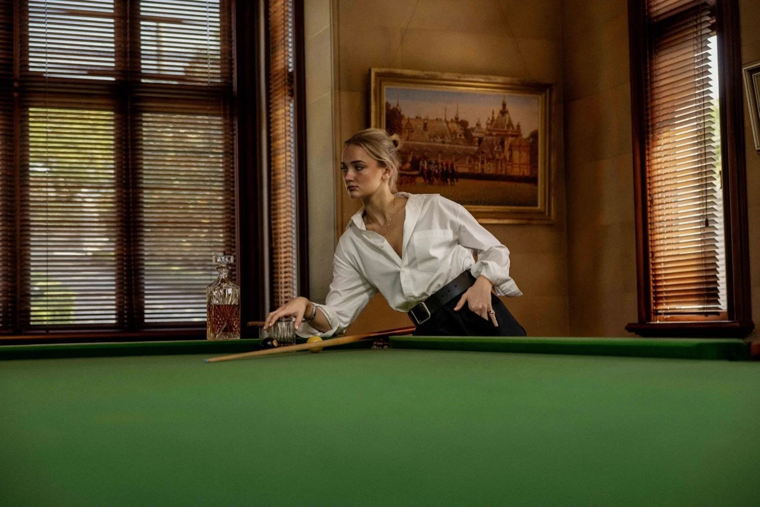A young woman in a white shirt and black pants is leaning over a pool table, preparing to take a shot. There is a glass of whiskey and a decanter on the table. The room has wooden window blinds and a framed painting of a historical building on the wa