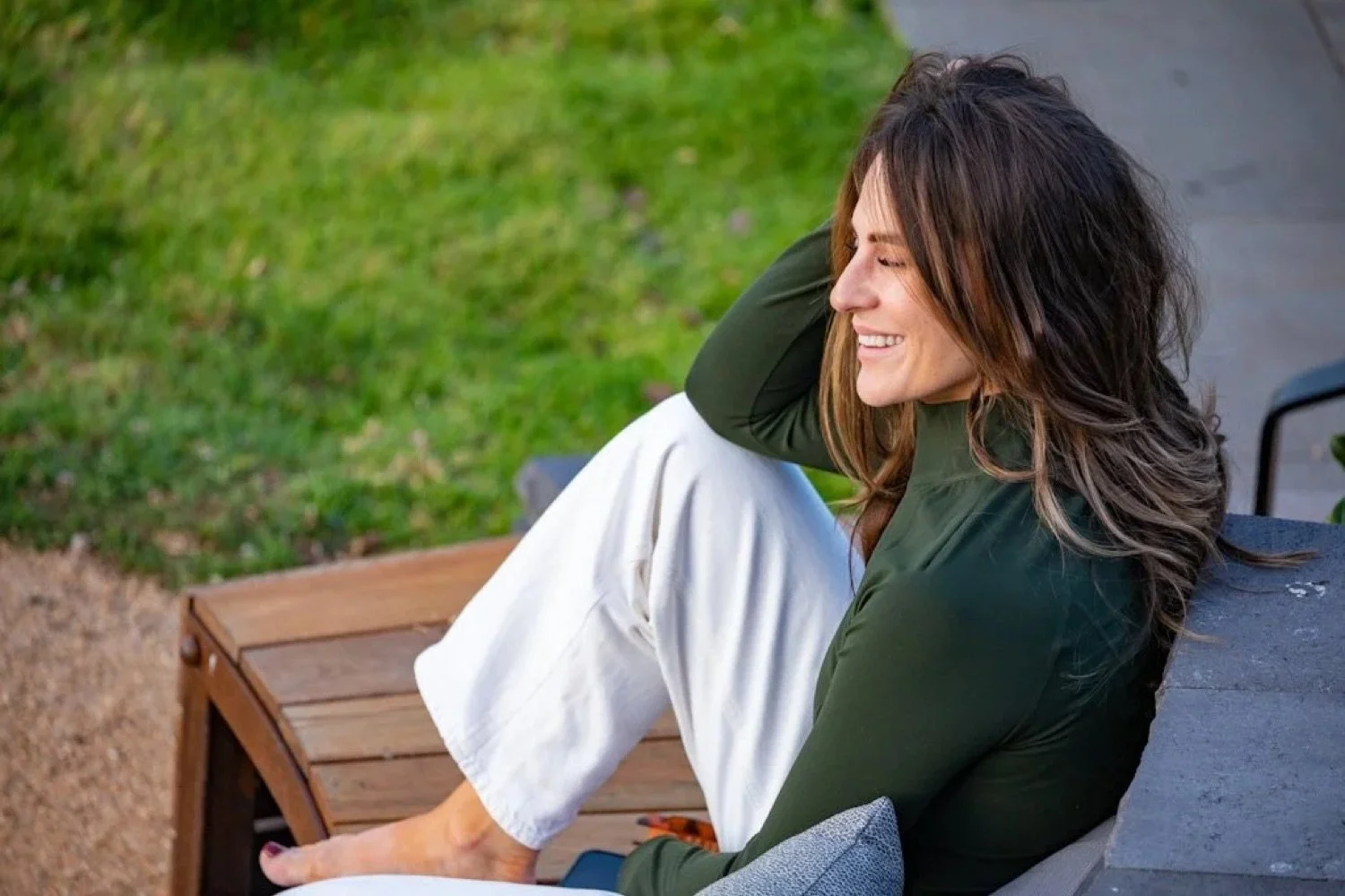 A woman with long brown hair sitting on a bench outside, smiling with her eyes closed, wearing a green jacket and white pants, in a park setting.