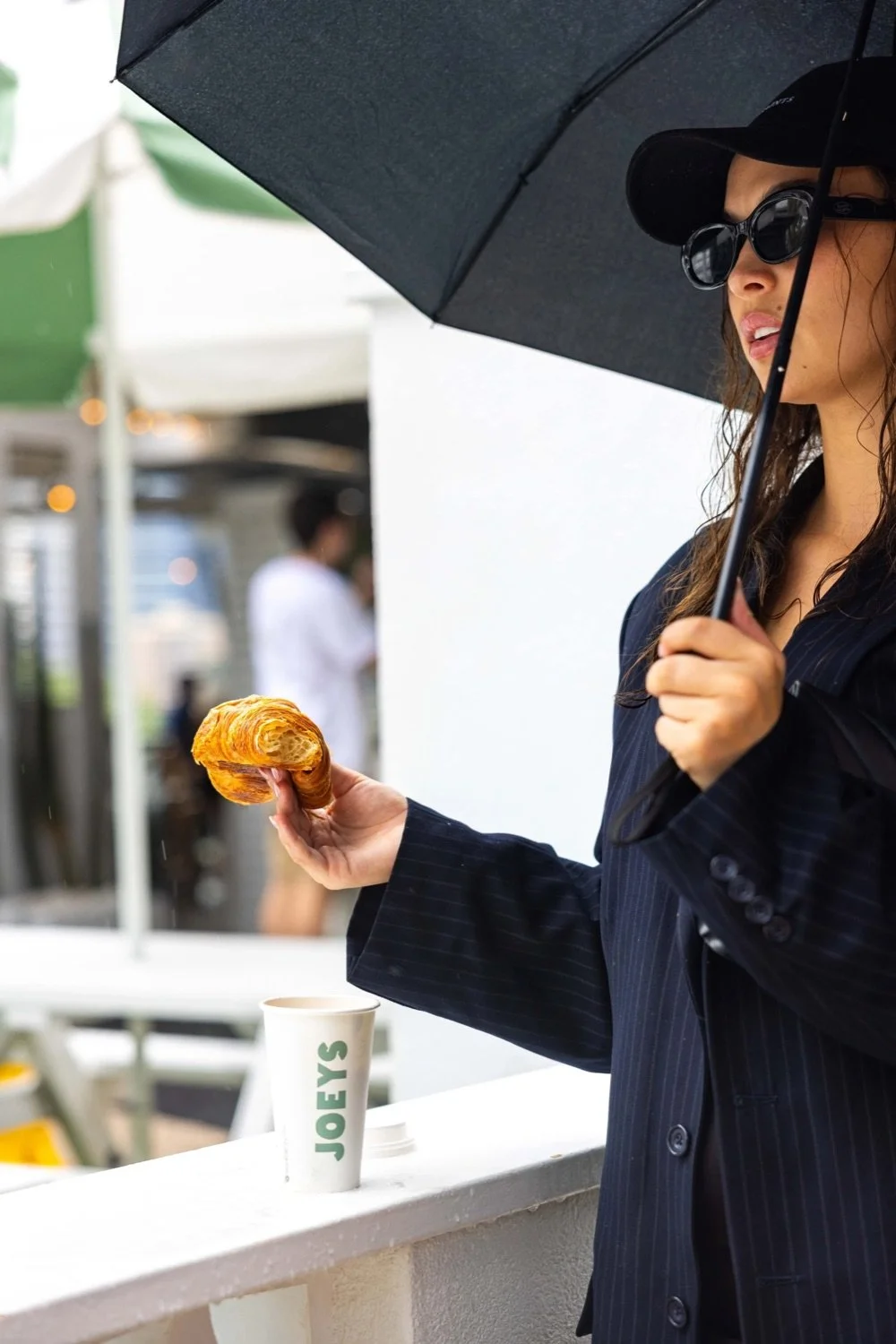 A woman holding a croissant under an umbrella near a Joe's coffee cup.