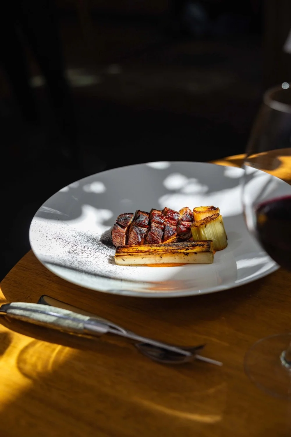 A white plate with slices of cooked beef, grilled leeks, and roasted green onions, placed on a wooden table with a glass of red wine nearby.